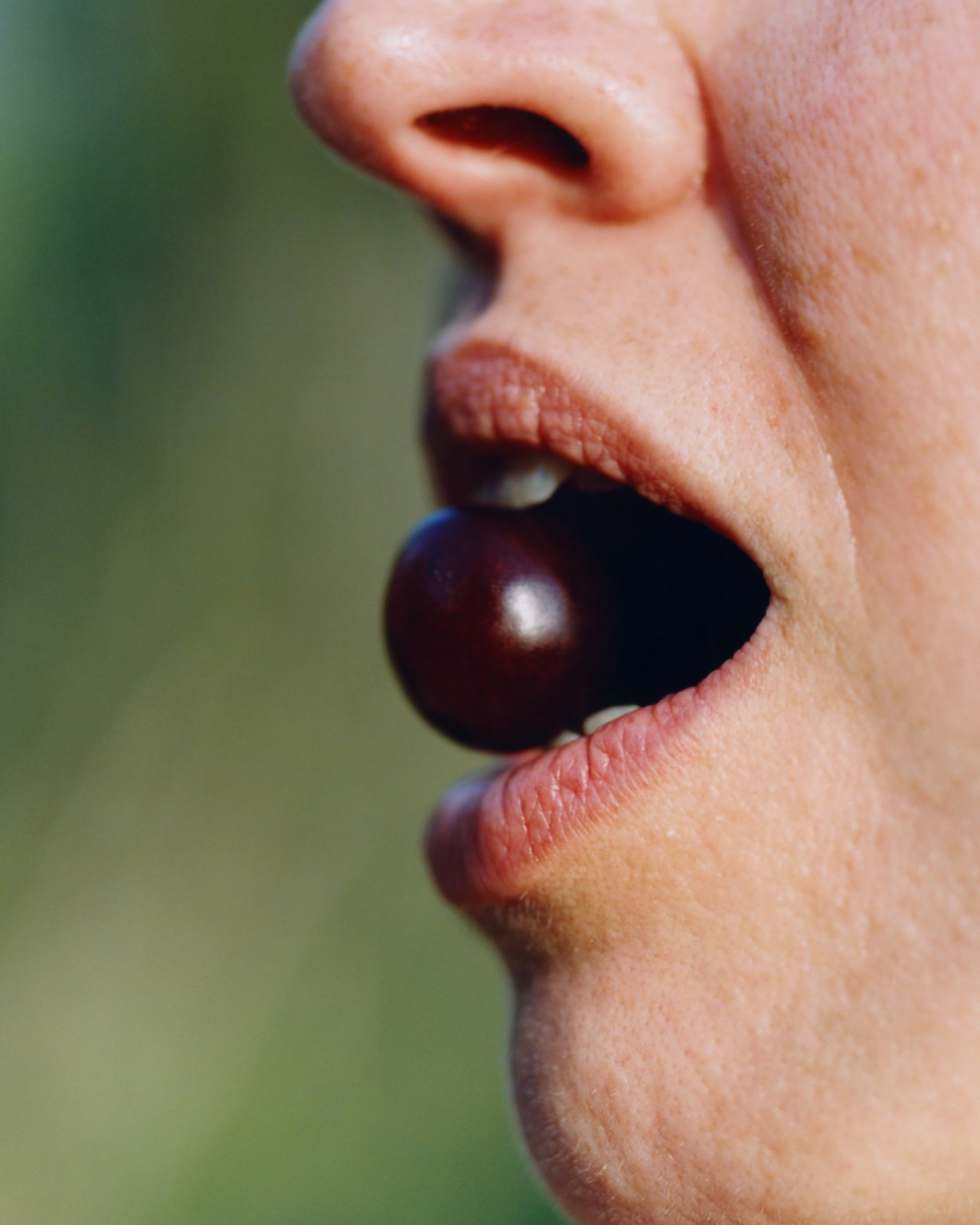 The profile of a person holding a red cherry between their teeth.