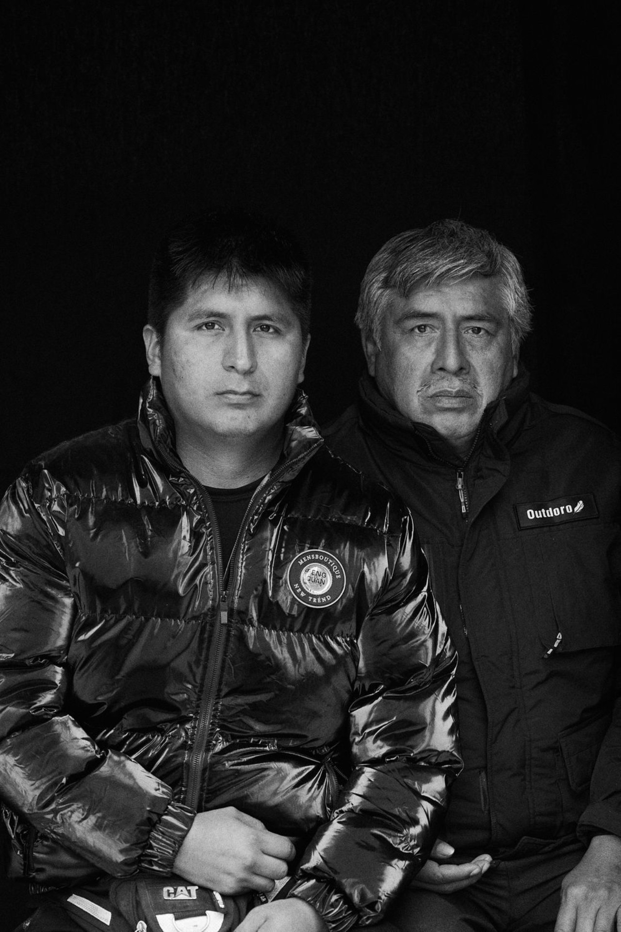 A black and white portrait of two Peruvian people sitting next to each other, looking seriously into the camera.