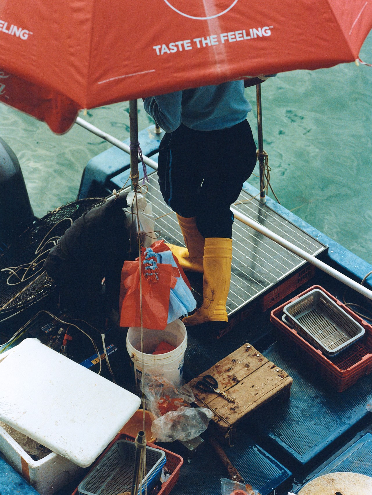 A person in yellow rain boots is covered by a large red umbrella while standing on a boat in a Hong Kong village.