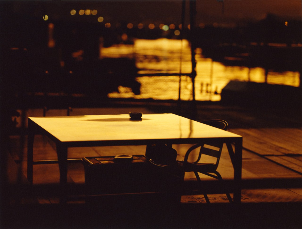 A table and chair are soak in the sun at dusk. The water in a Hong Kong village is in the background.