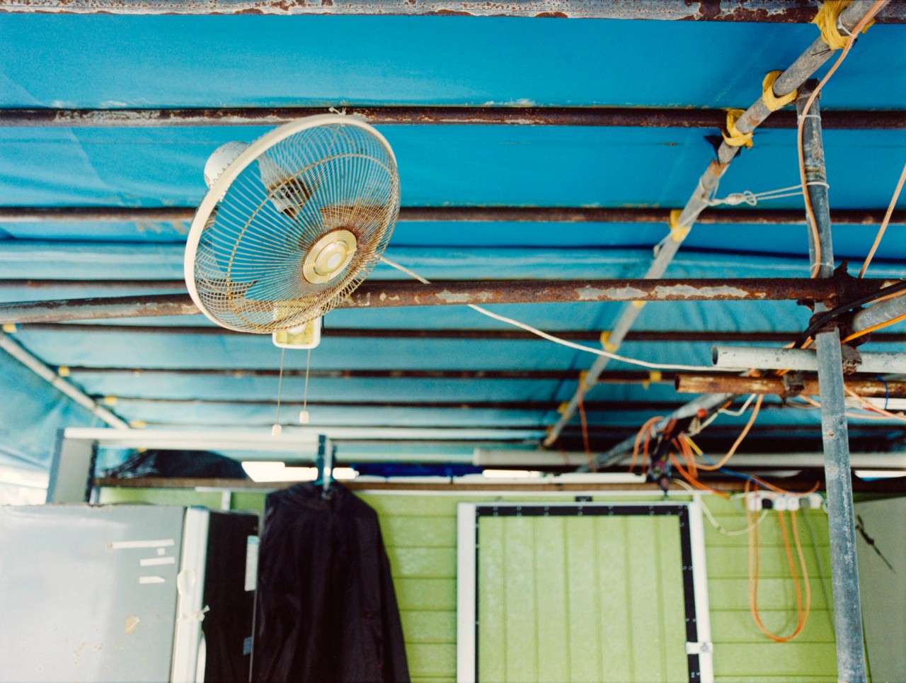 A fan hangs on the roof of a blue shack.