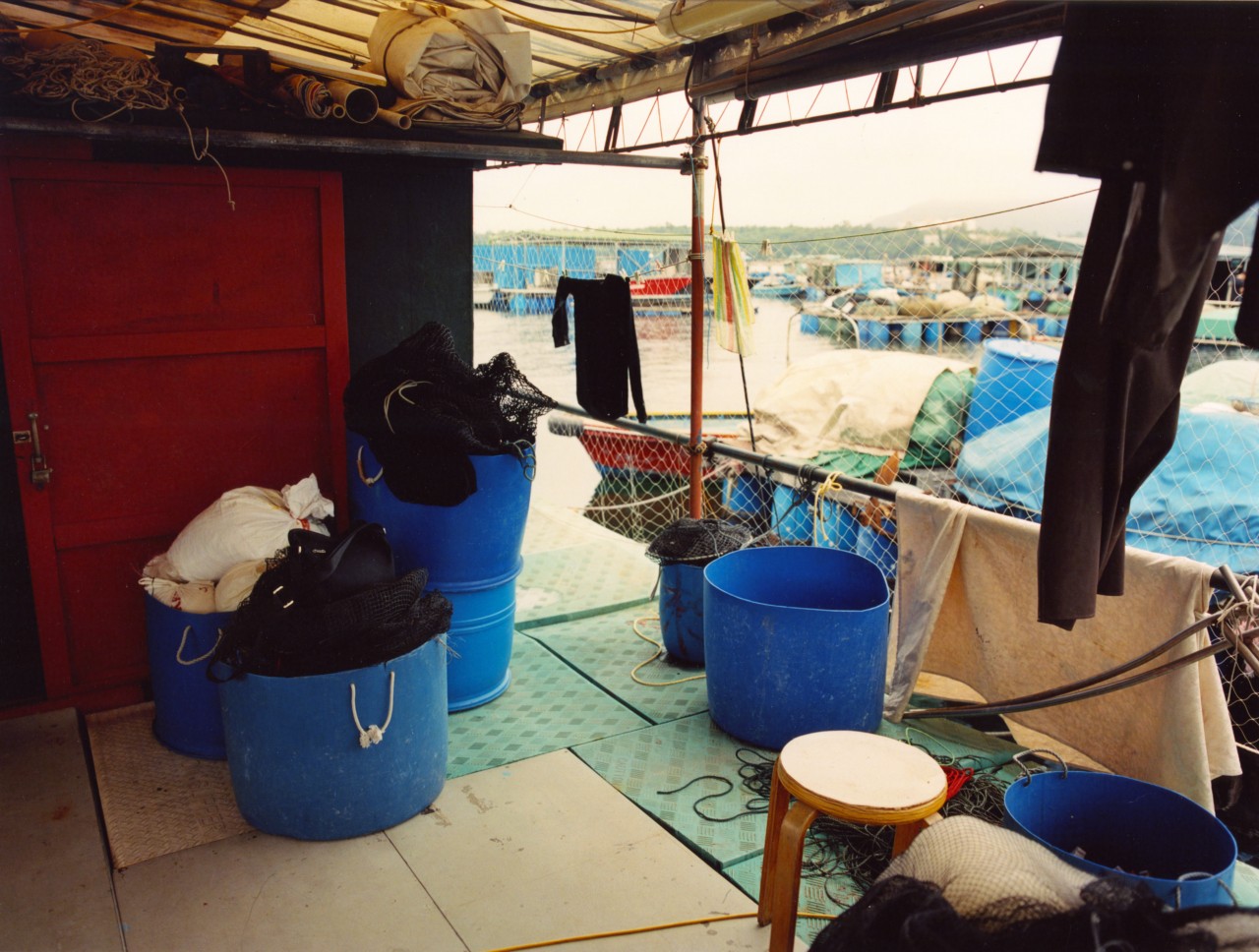 The inside of a shack shows large blue buckets and opens up to the outside of a fishing village.