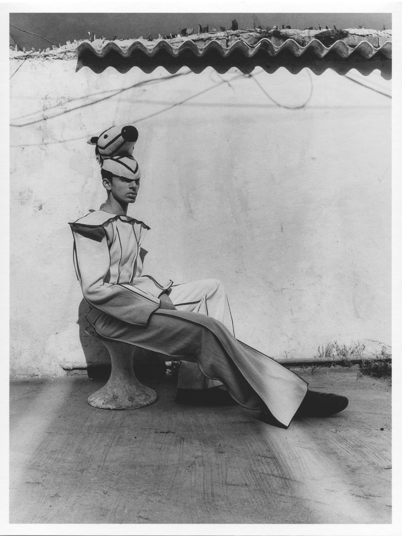 A model wearing pantsuit and a horse hat poses while sitting down in front of a wall.