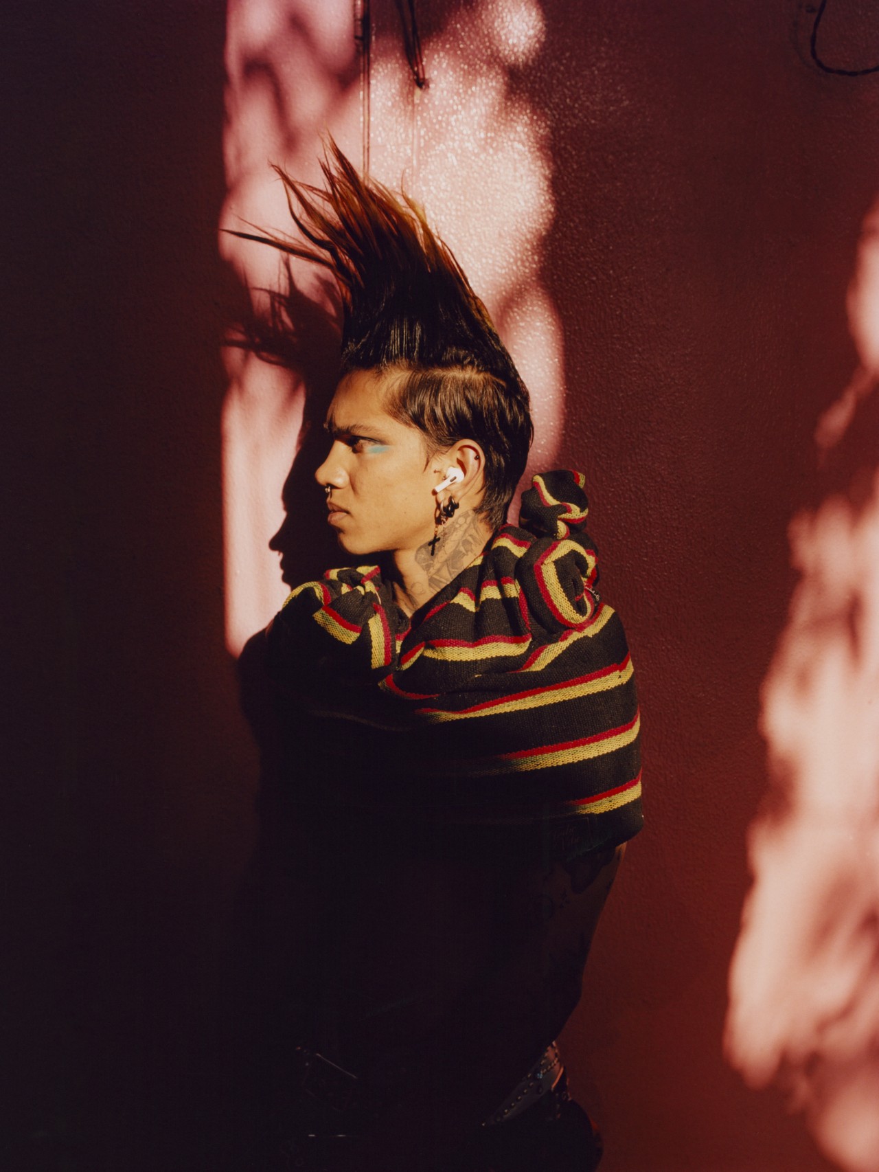 A model with a tall mohawk faces the side. They are standing in front of a pink wall, halfway hidden in the shadows.