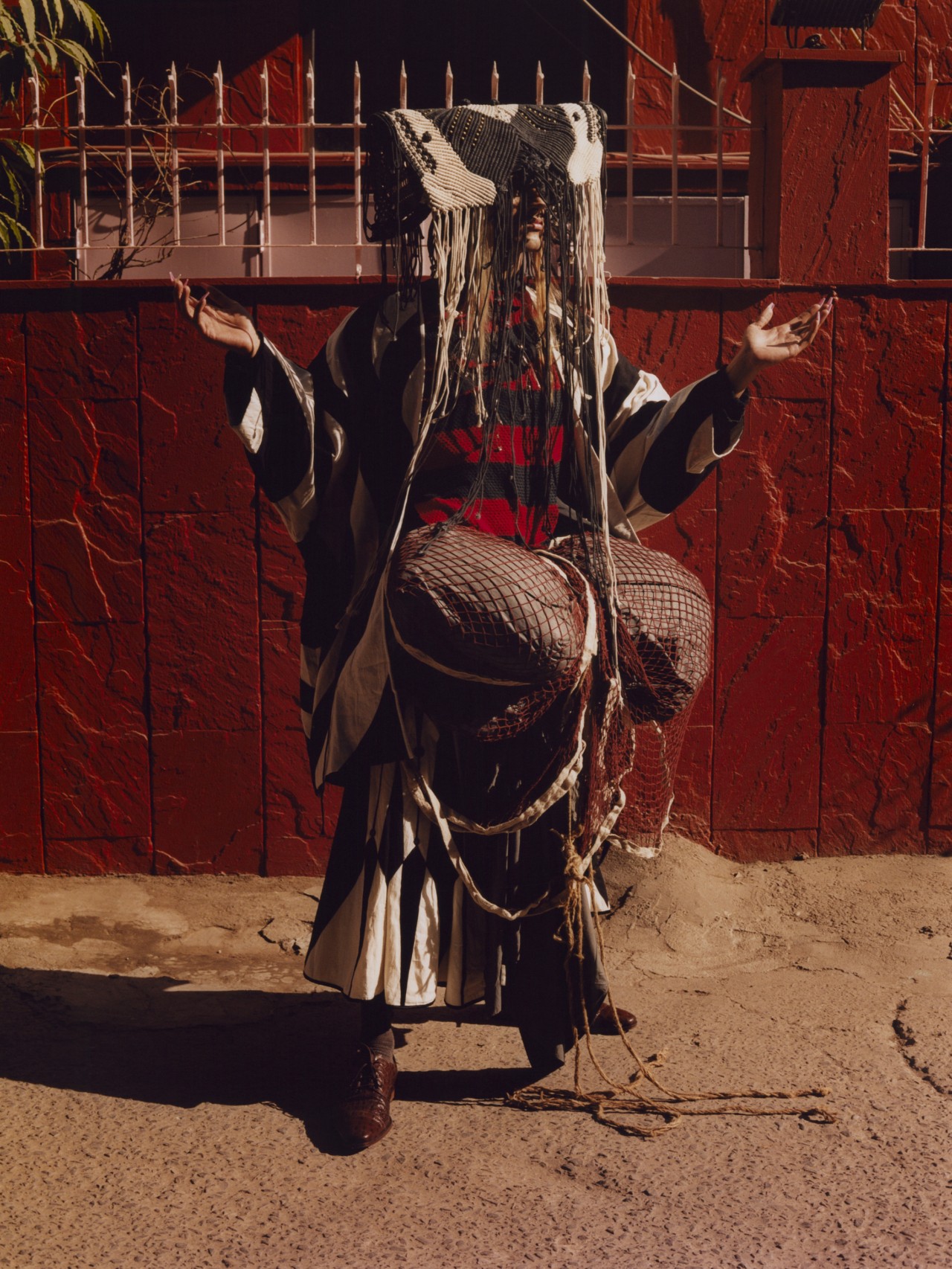 A model holds their hands up while wearing an intricate garment. They're standing in front of a red building.