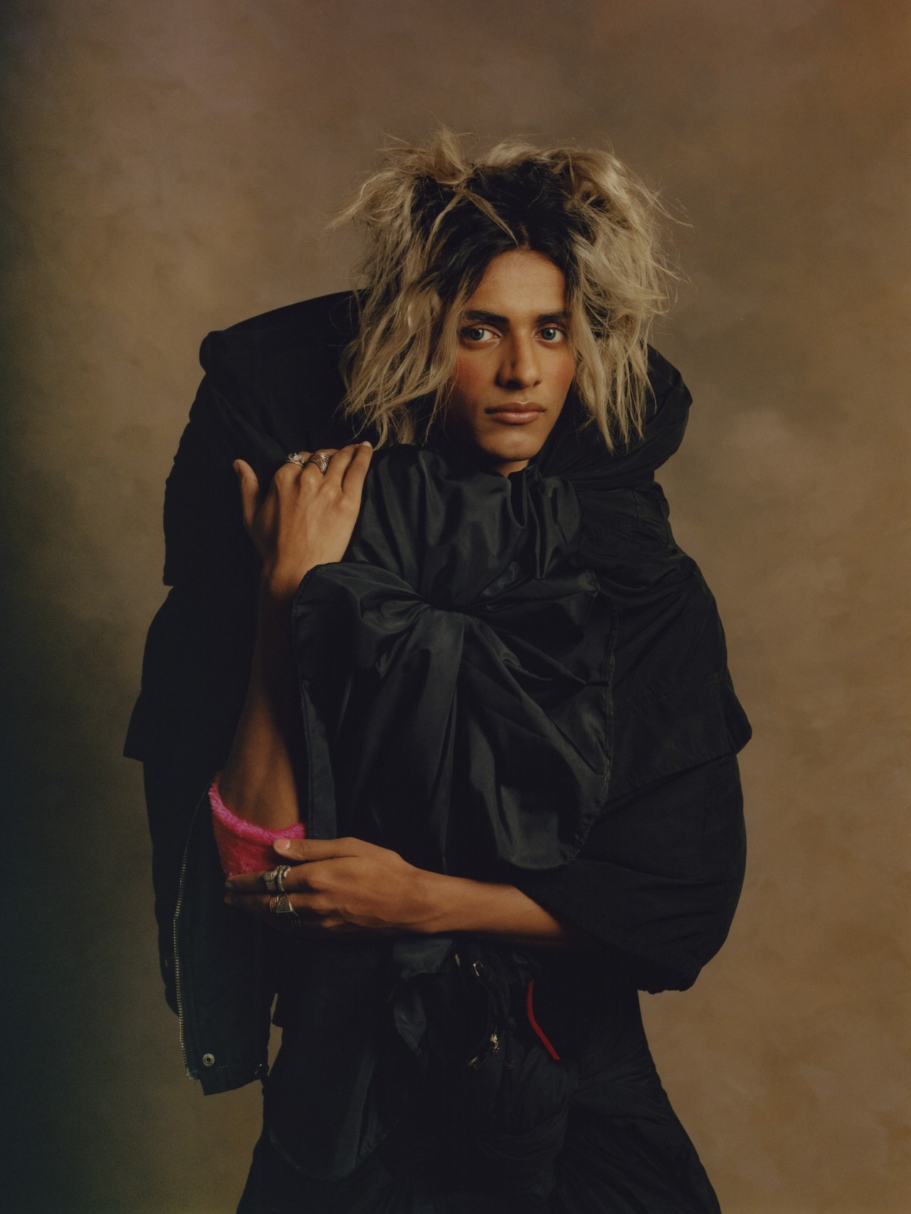 A model with blonde hair, wearing a heavy black garment looks into the camera.
