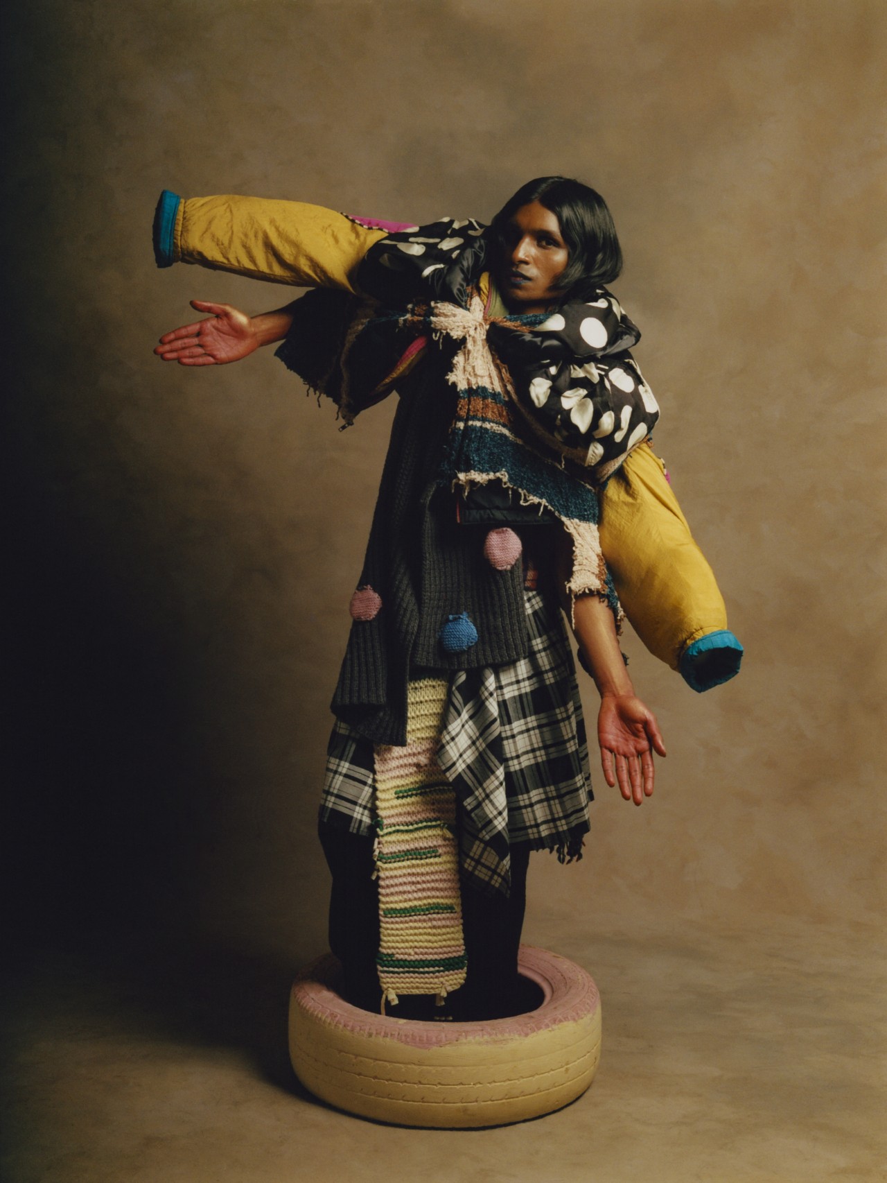 A model wearing layers of clothes stands inside a tan colored tire while holding out their arms.