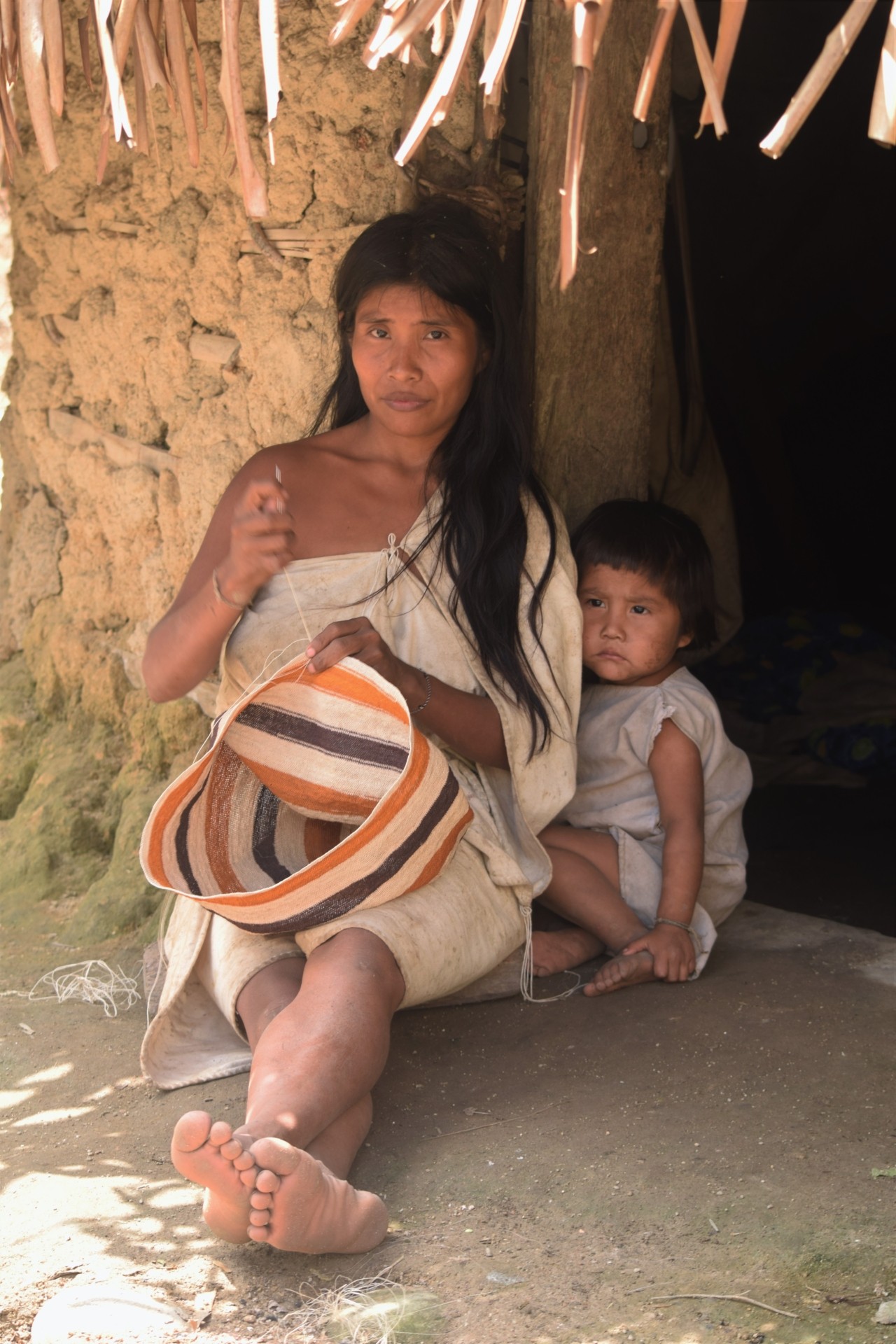 A Kogi woman sits while weaving a basket. A Kogi child sits right behind her.