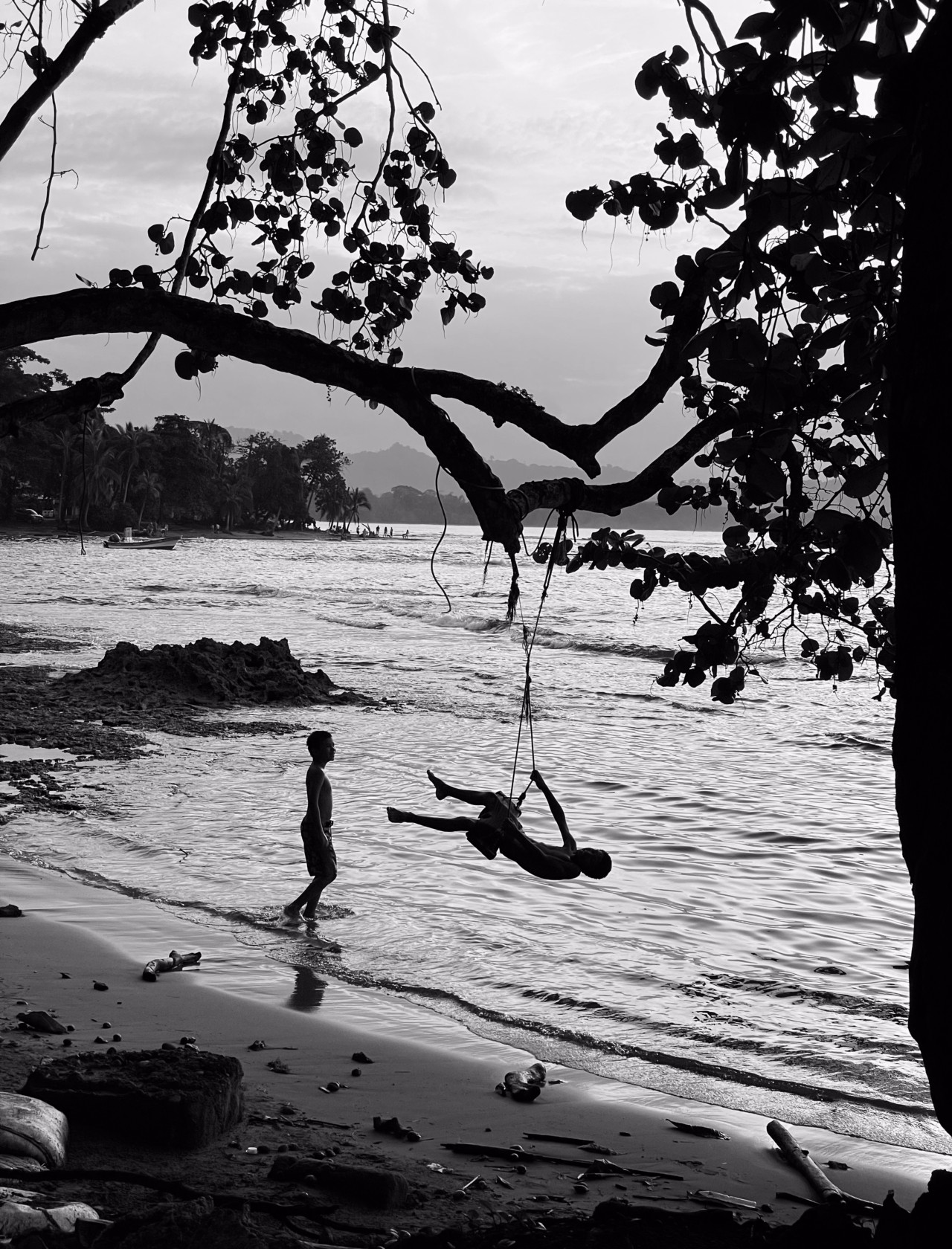 Two children play on a beach in Puerto Viejo. One of the children is swinging from a home made swing that is attached to a tree branch above him.