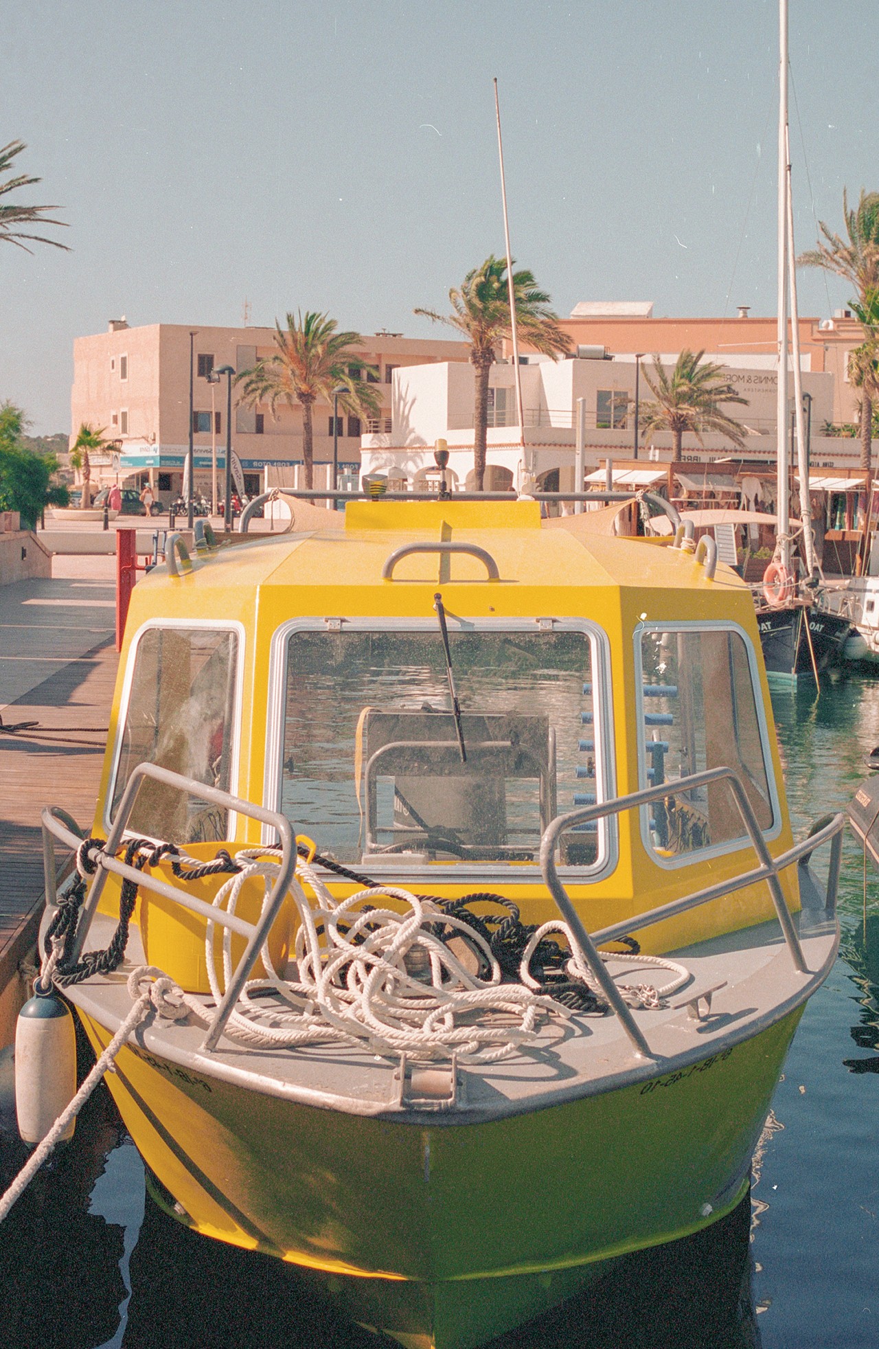 A yellow and white boat is docked in the sea. Behind it are palm trees and buildings.