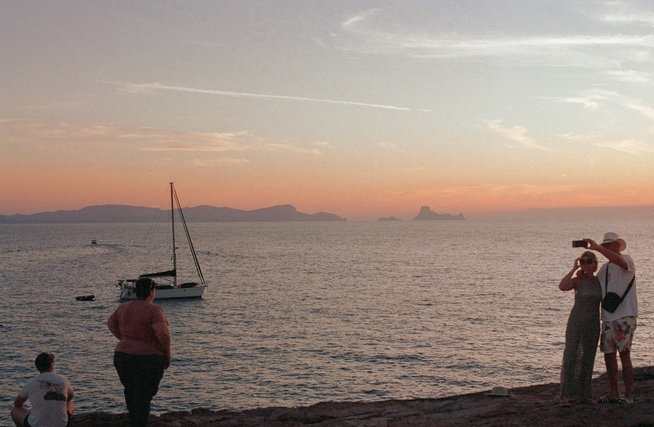 In Spain, people stand along the coast during sunset in front of the vast sea.