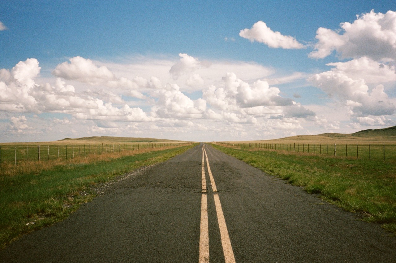A road surrounded by green grass with a cloudy sky above, in Clayton, NM