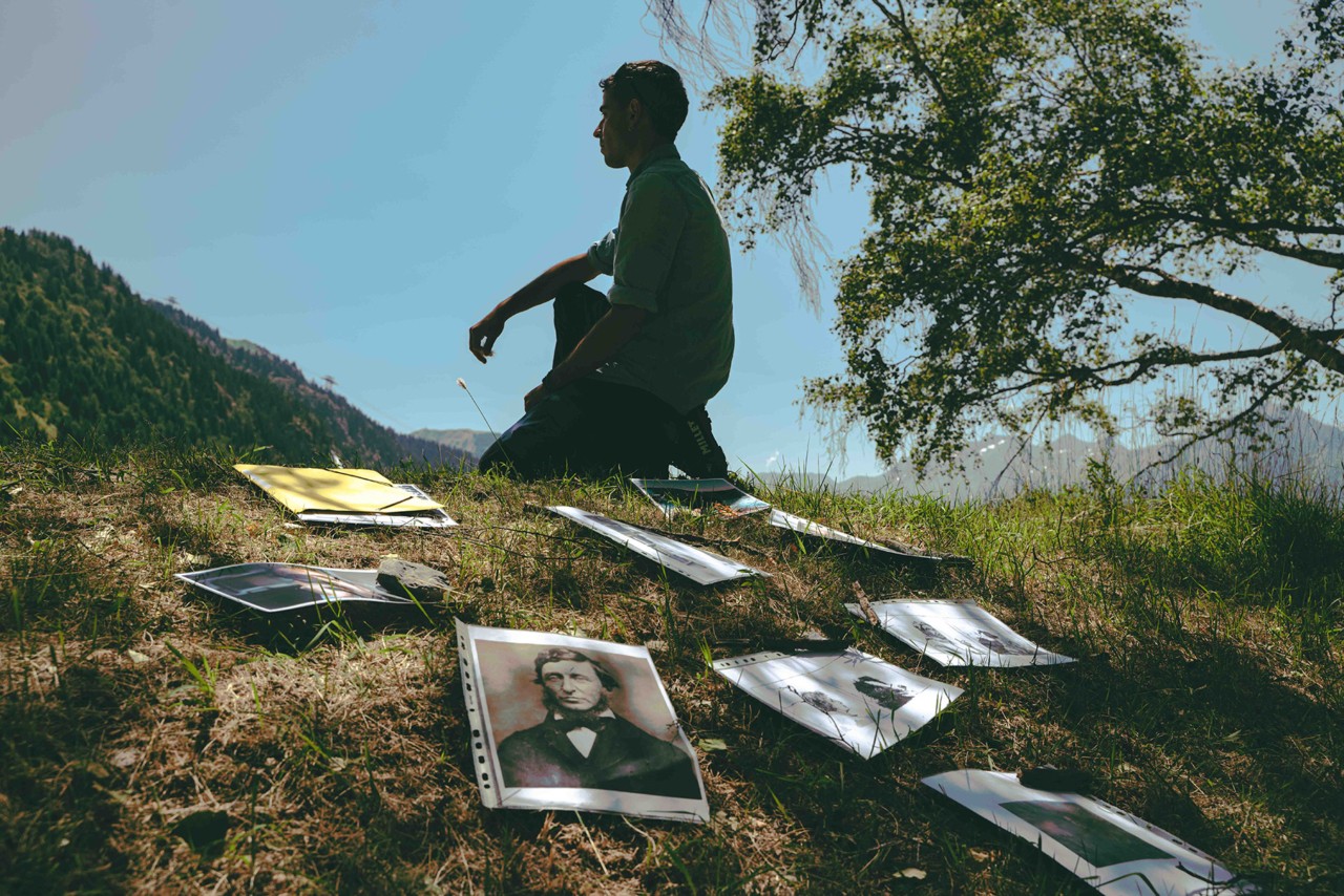 A person sits on a grassy mountain top looking in the distance. Next to them are old photographs scattered.