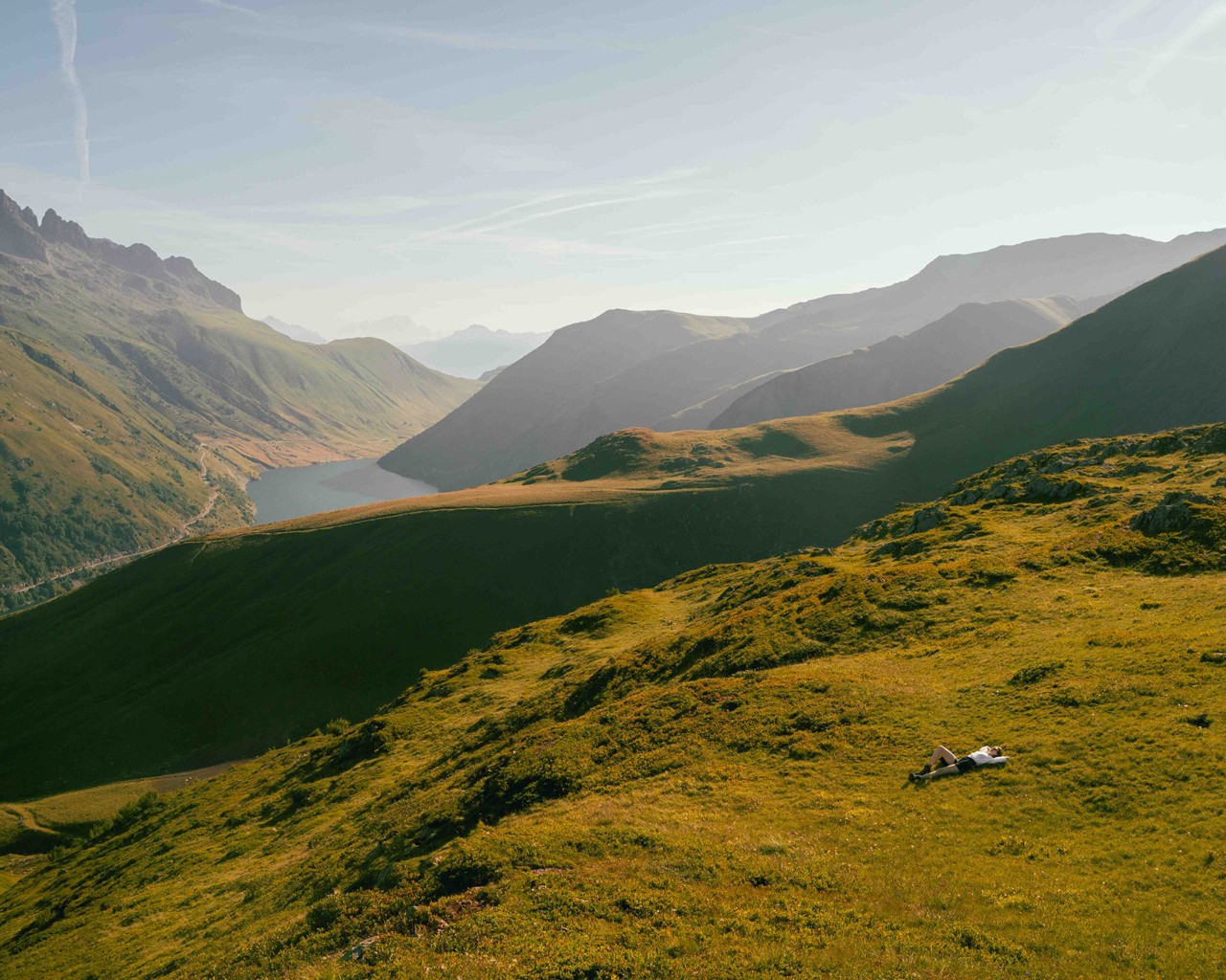 A person lies on vast green mountains that descend into a valley filled with water.