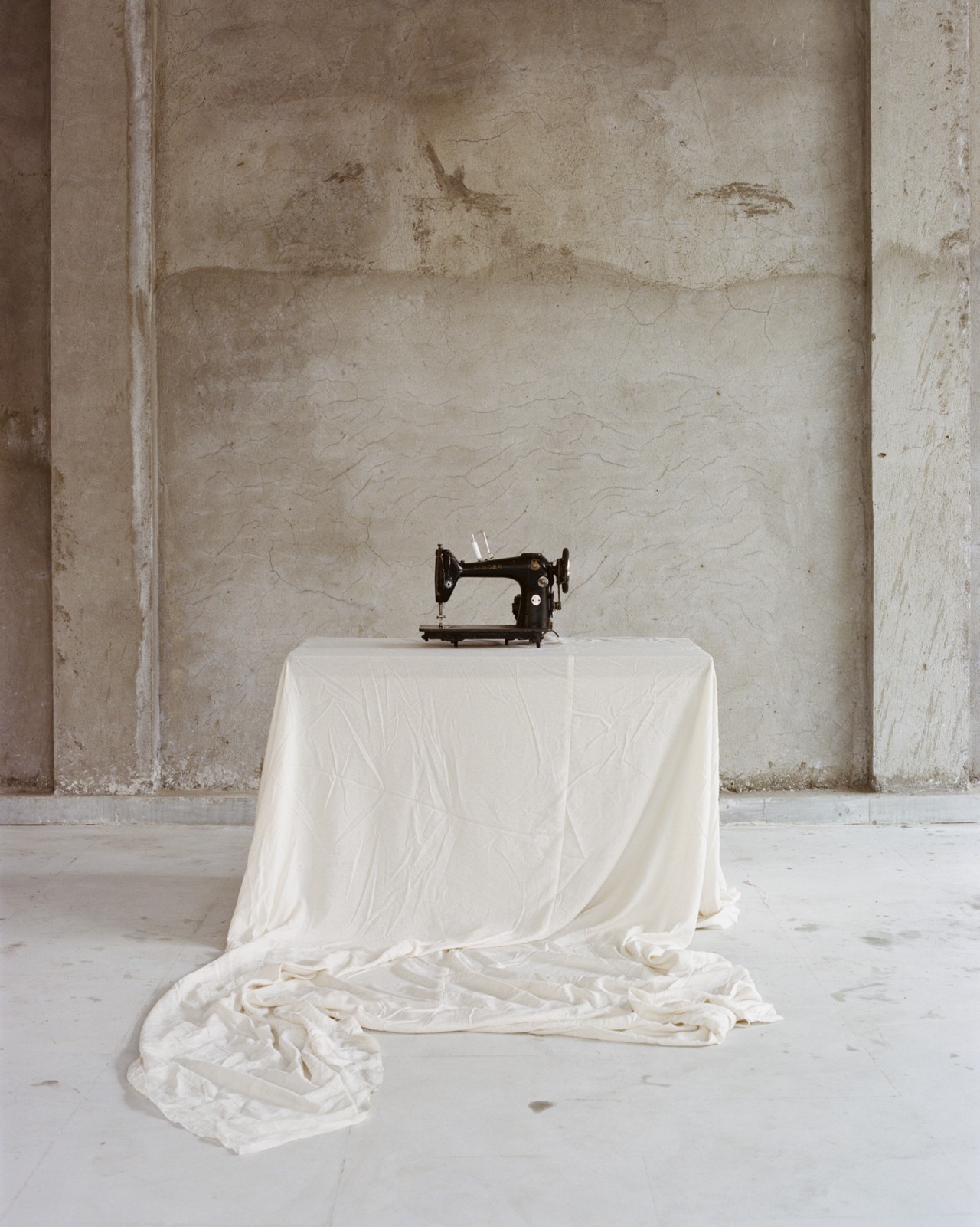 A black sewing machine rests on a table draped with a white sheet in an empty concrete room.