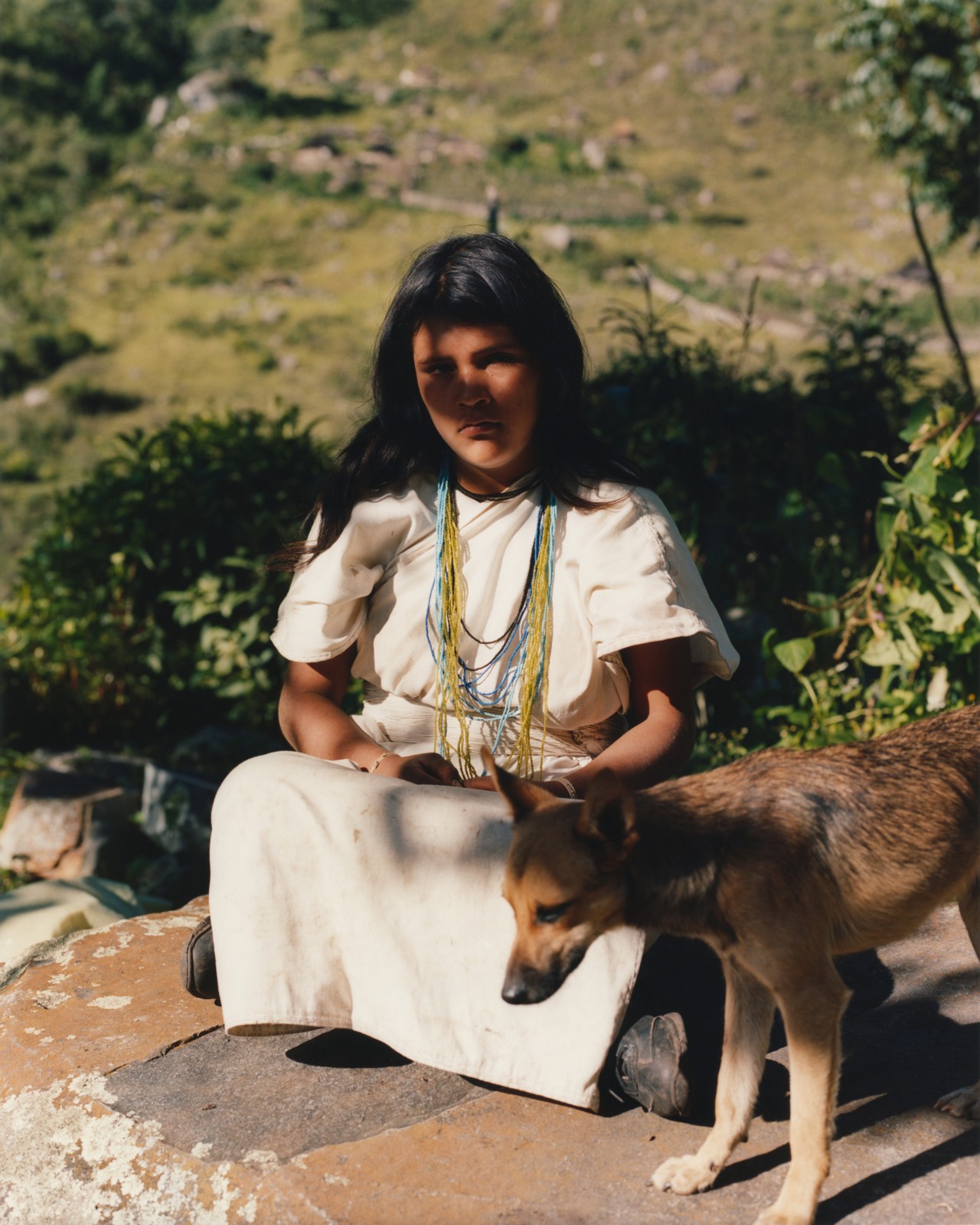 A young Kogi person sits next to a dog among the rocky terrain of Colombia.