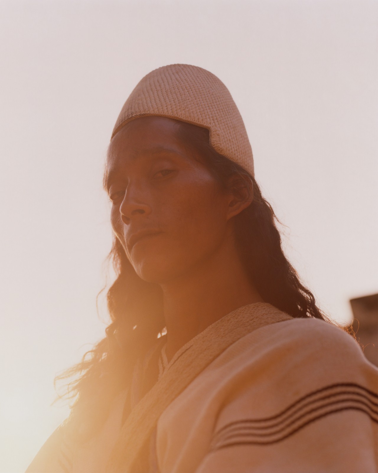 A Kogi person dressed in traditional garb peers down at the camera with the sun beaming behind them.
