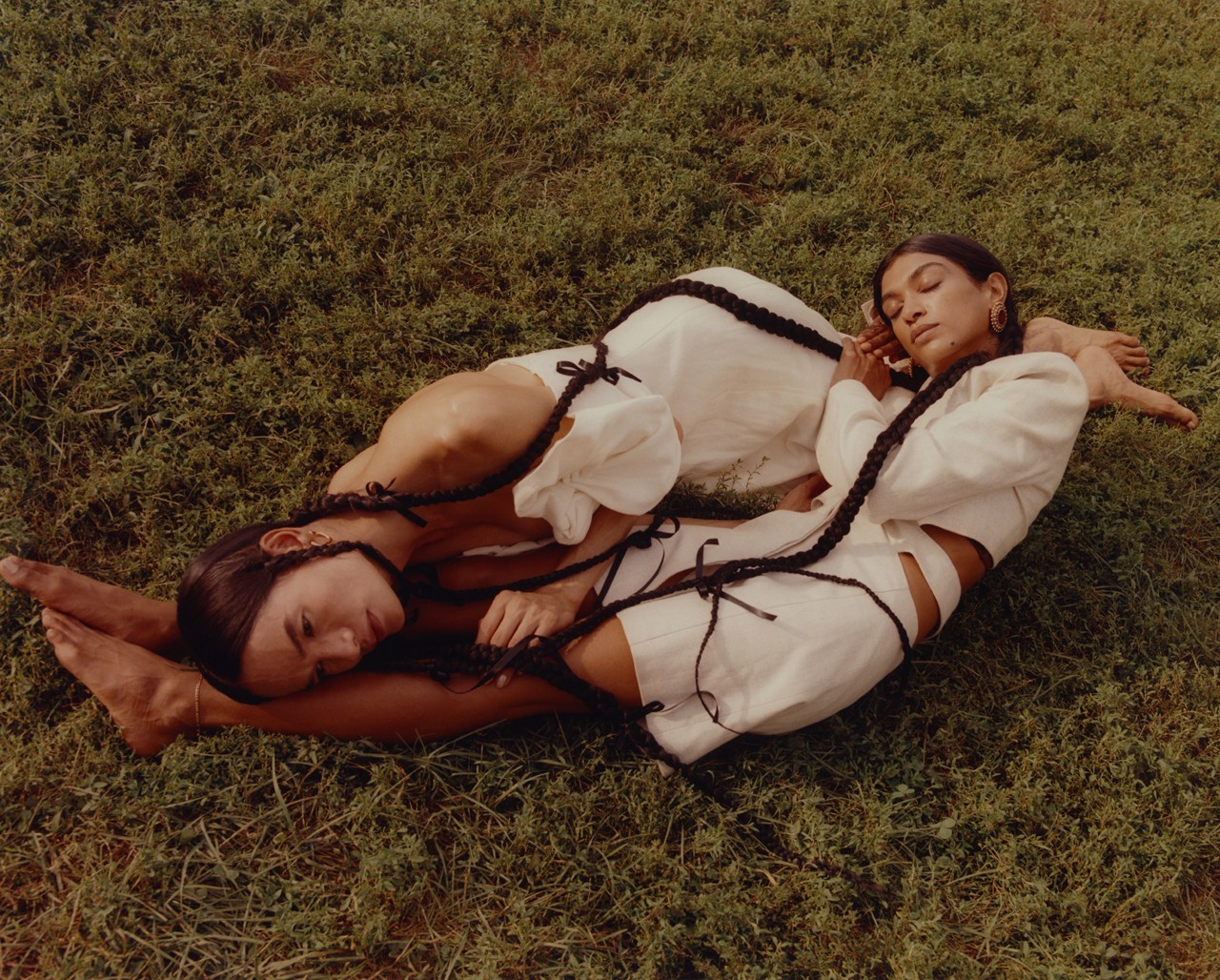 Two models with long braids, wearing white linen fabric outfits lay next to each other in a grassy field.