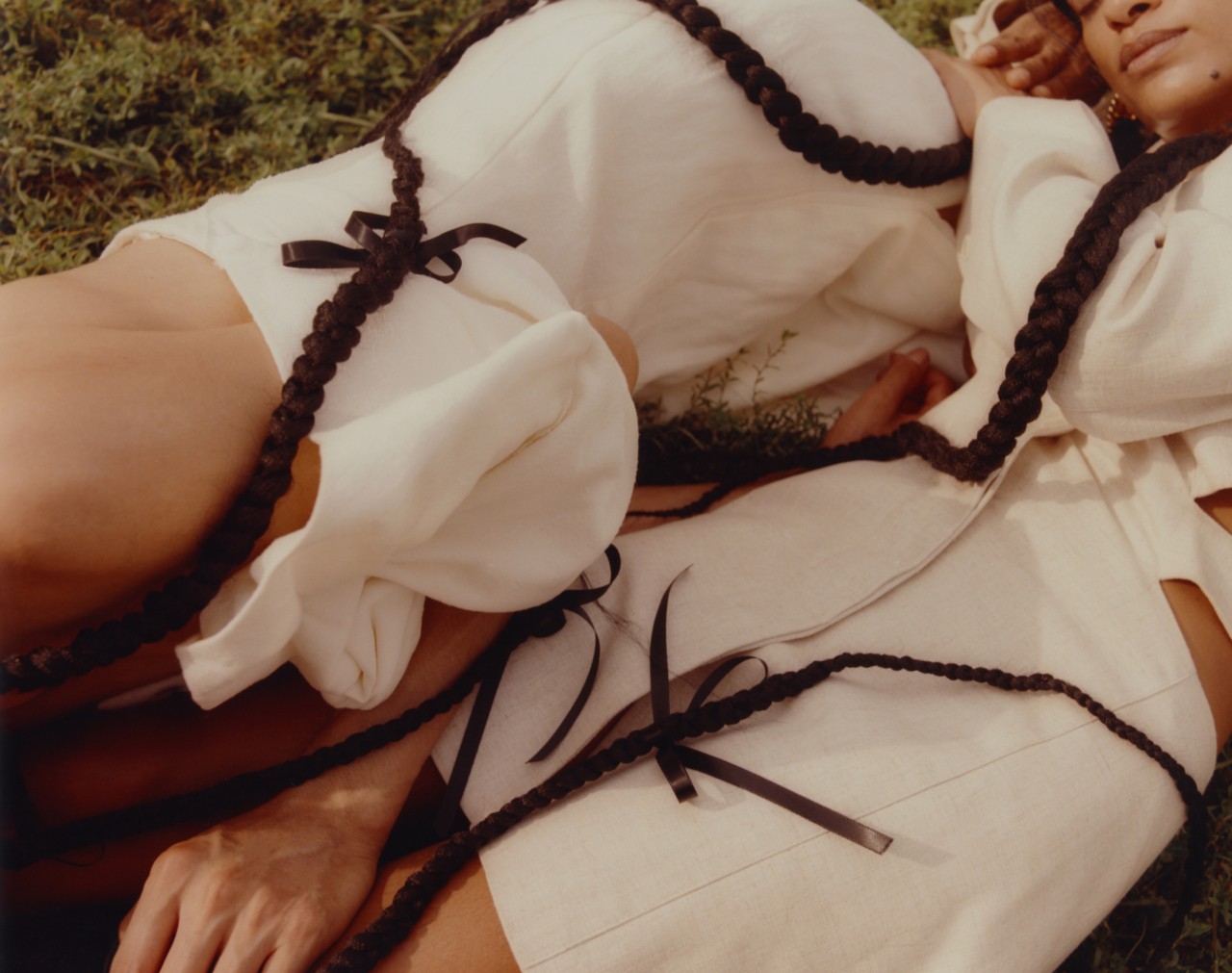 Two models with long brown braids wearing white linen fabric lay next to each other on the grassy ground.