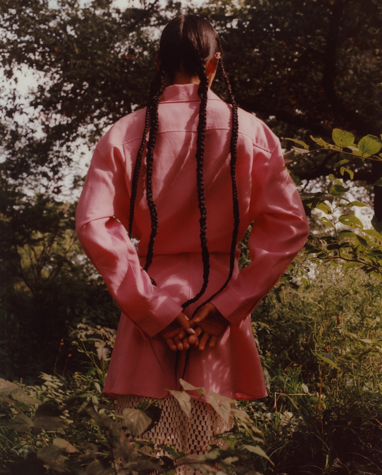 A model with long braids is wearing a pink linen jacket and skirt and showing their back to the camera while standing in a forest.