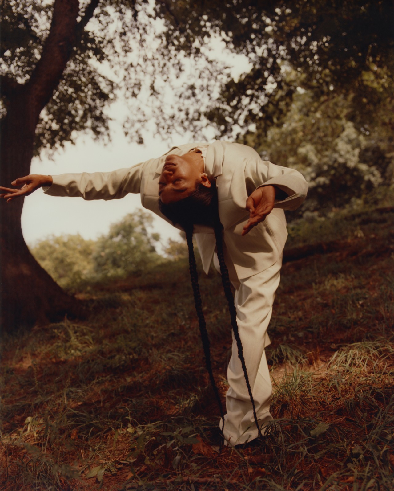 A model wearing a beige linen suit closes their eyes while leaning backwards in the middle of a forest.