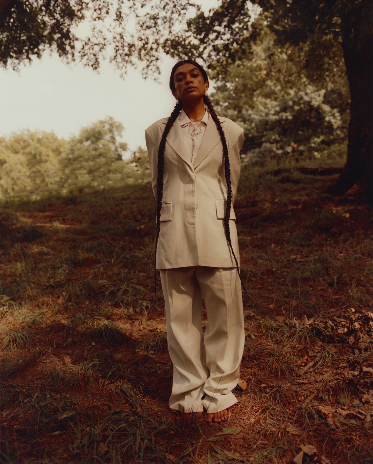 A model wearing a large beige linen suit stands in the middle of the forest, looking straight ahead with their hands behind their back.