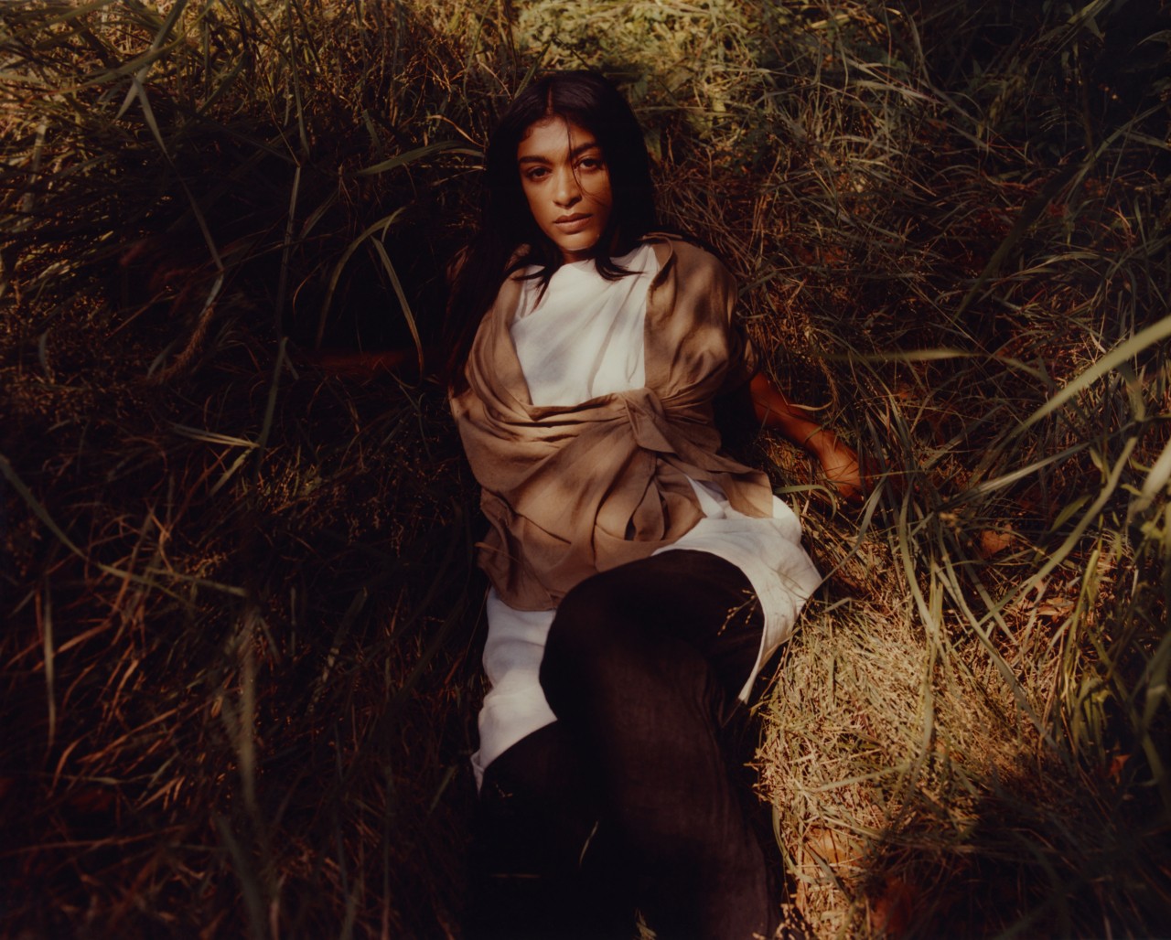A model wearing white, brown and black linen fabric looks up at the camera while laying down in a field of weeds.
