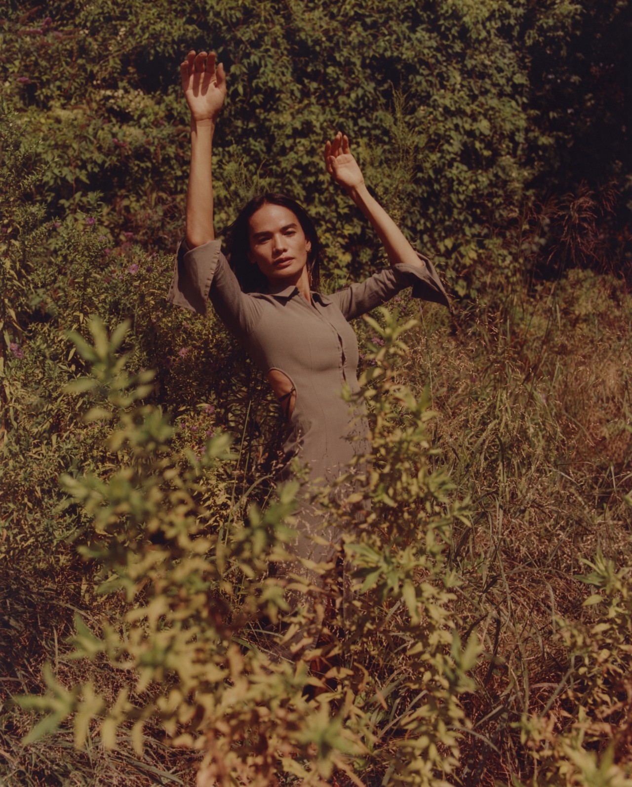 A model standing amongst tall leaves in a forest is holding their hands up while looking toward the camera. They are wearing a taupe linen fabric outfit.