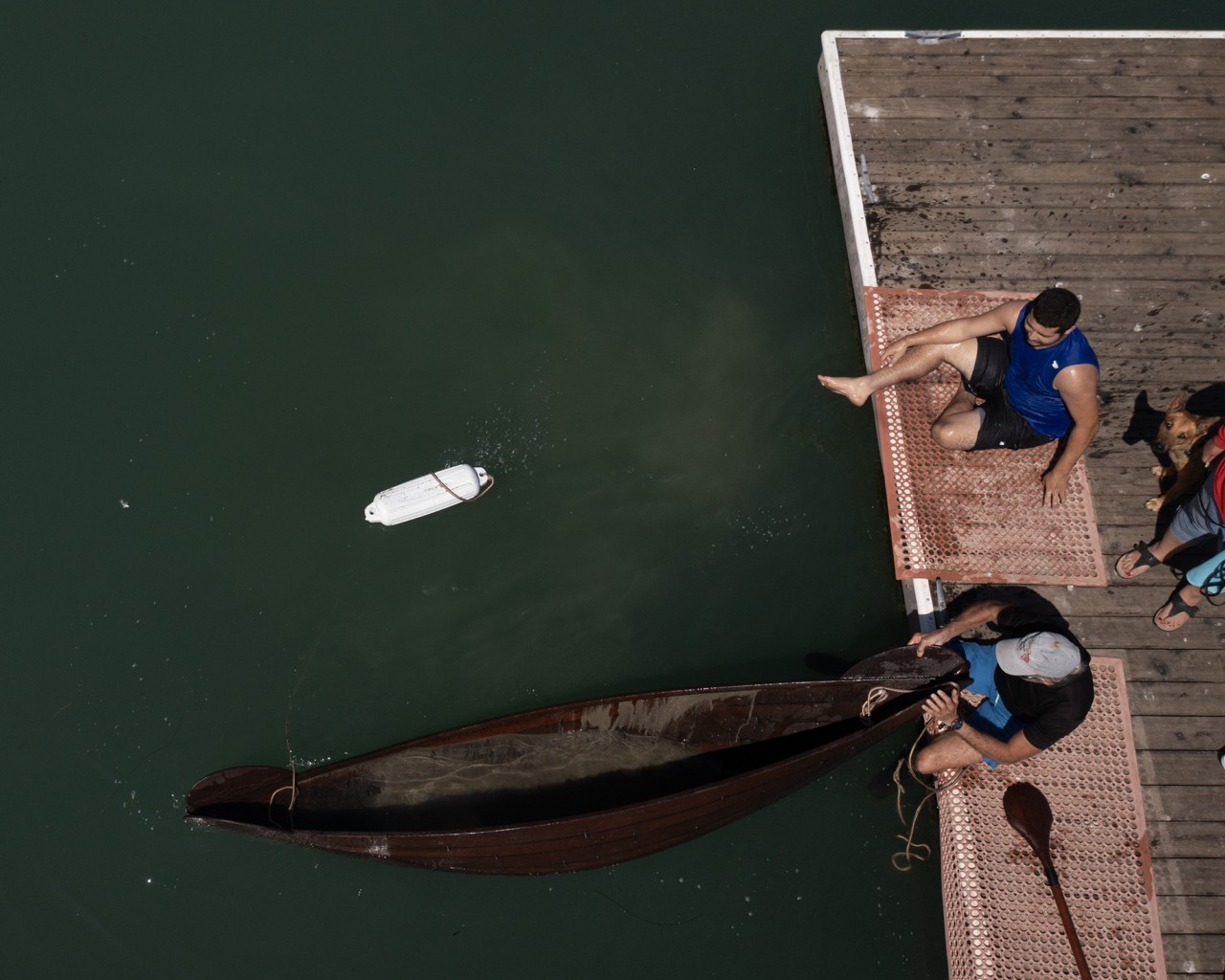 Two people sit at the edge of a dock. One is grabbing on to a canoe that it floating in the water.