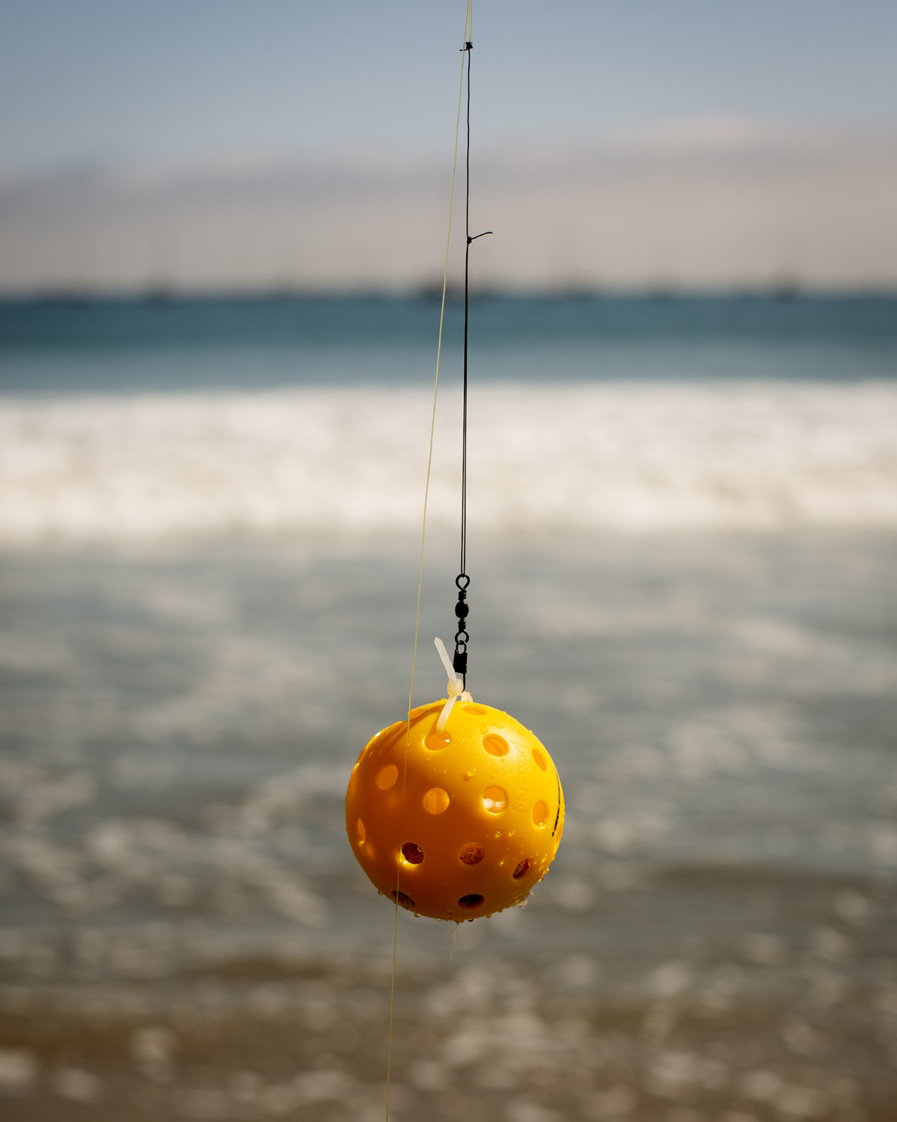 A fishing line holds a small, plastic orange ball on the hook. The waves of the ocean crash in the background.