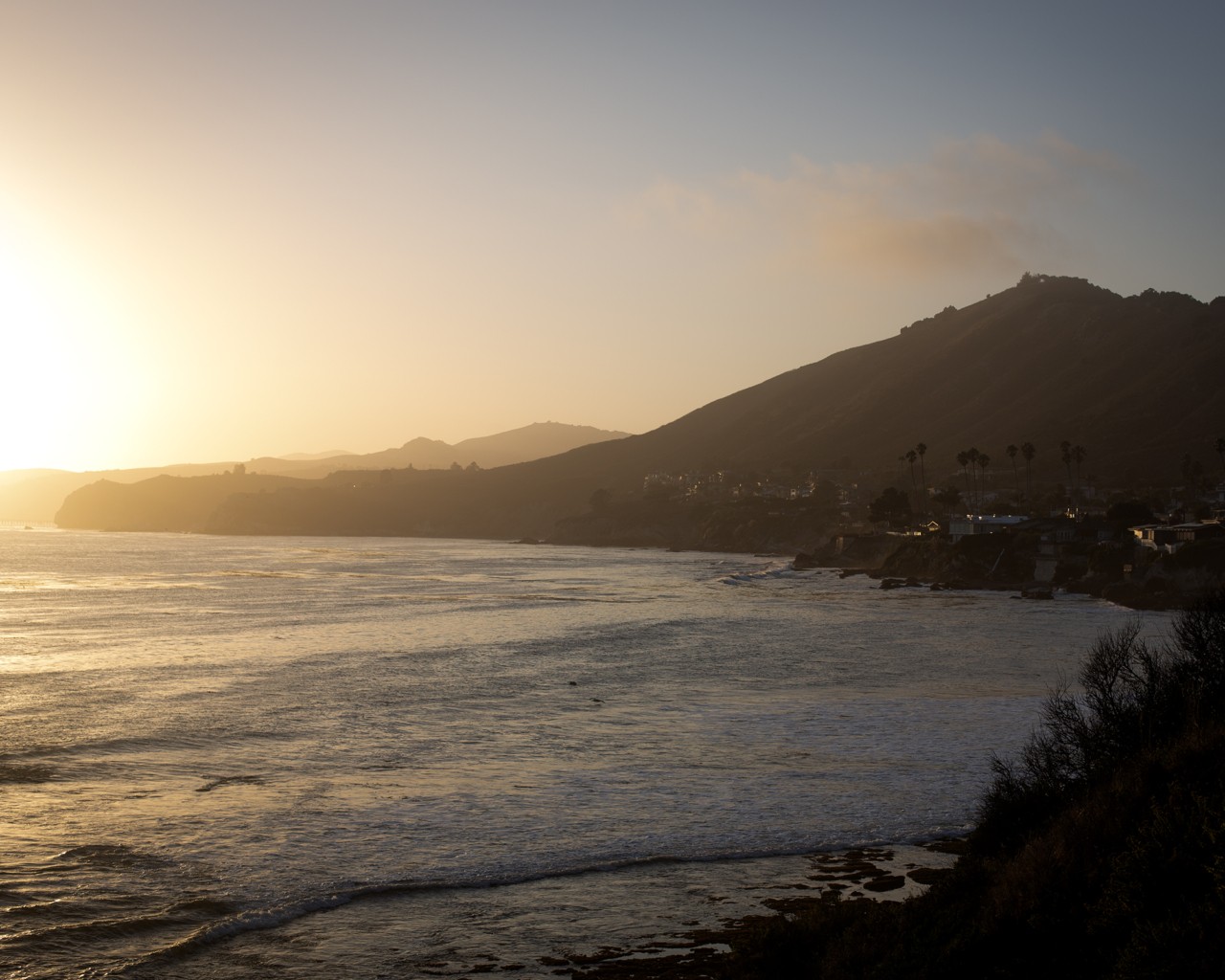 The sun sets over the ocean with mountains in the distance.