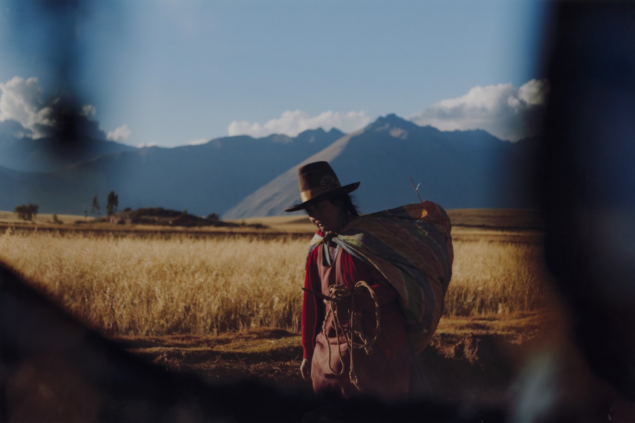 A rancher carries a large bag on his back as he walks along the road with the mountains behind him.