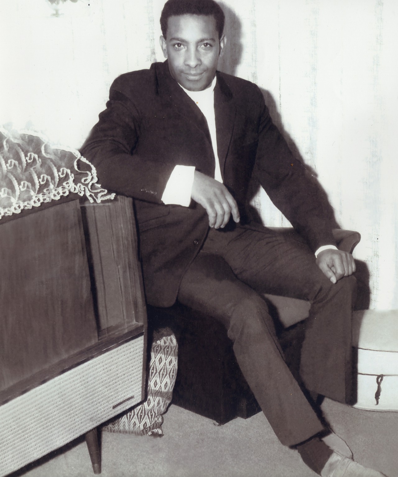 A vintage photograph of a man wearing a suit sitting in a chair in a bedroom.