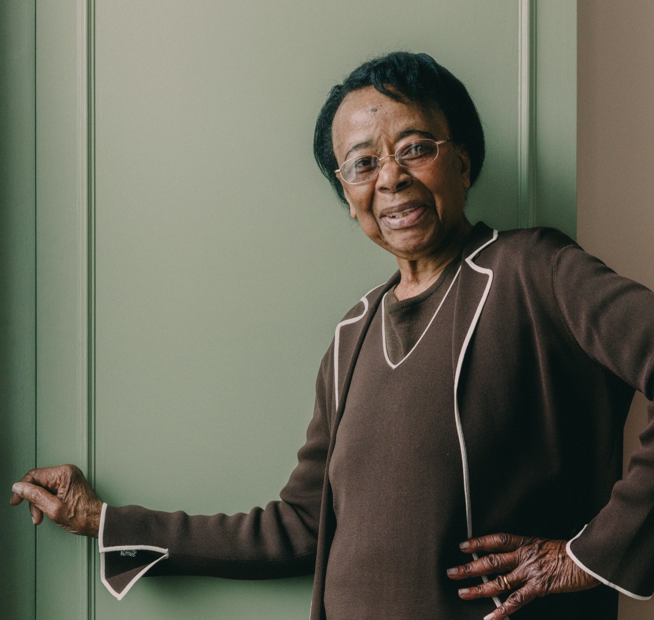 An older woman wearing a brown sweater smiles while leaning against a green door.