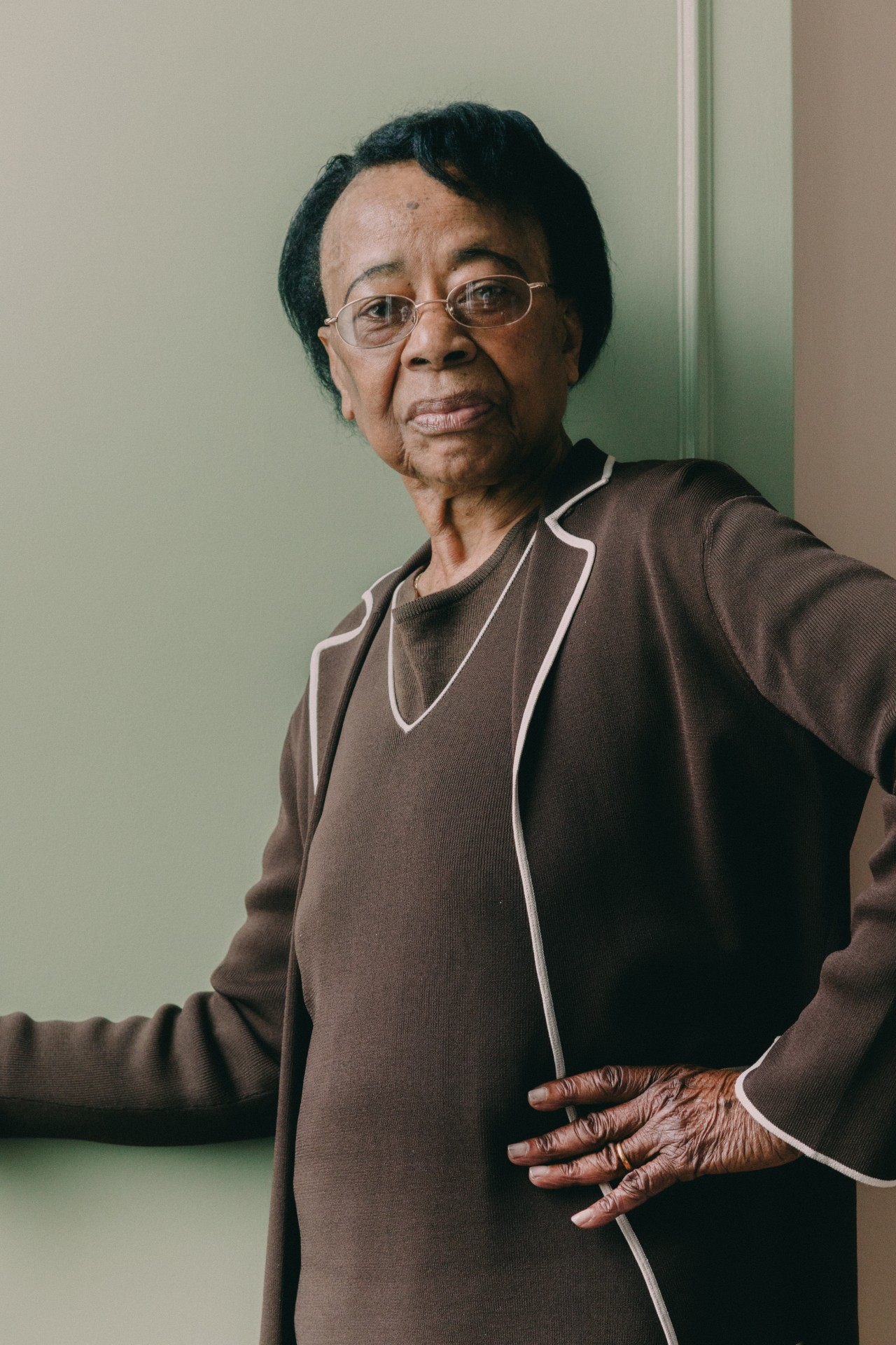 An older woman wearing a brown sweater and glasses poses with her hand on her hip in front of a green door.