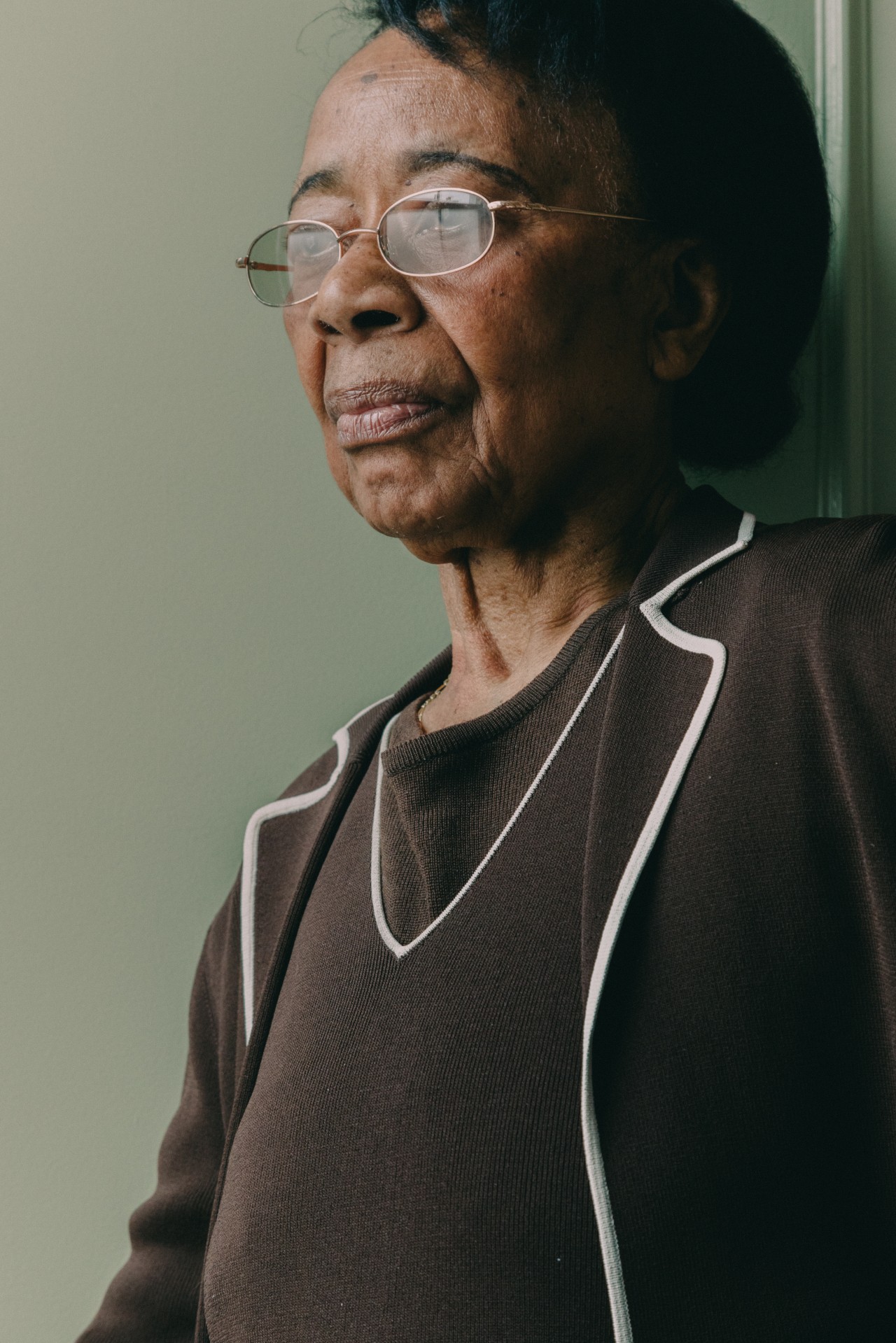 An older woman wearing a brown sweater and glasses looks into the distance.