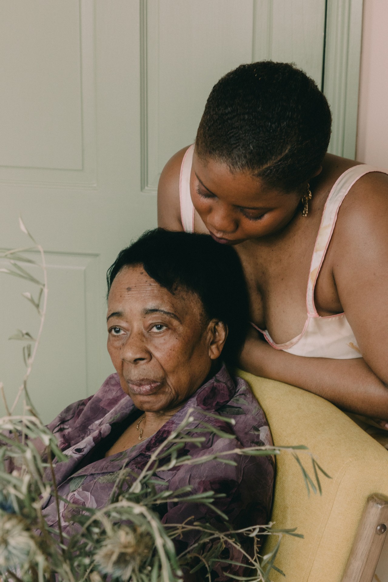 A younger woman rests her head on an older woman who is sitting in a yellow chair inside a home.