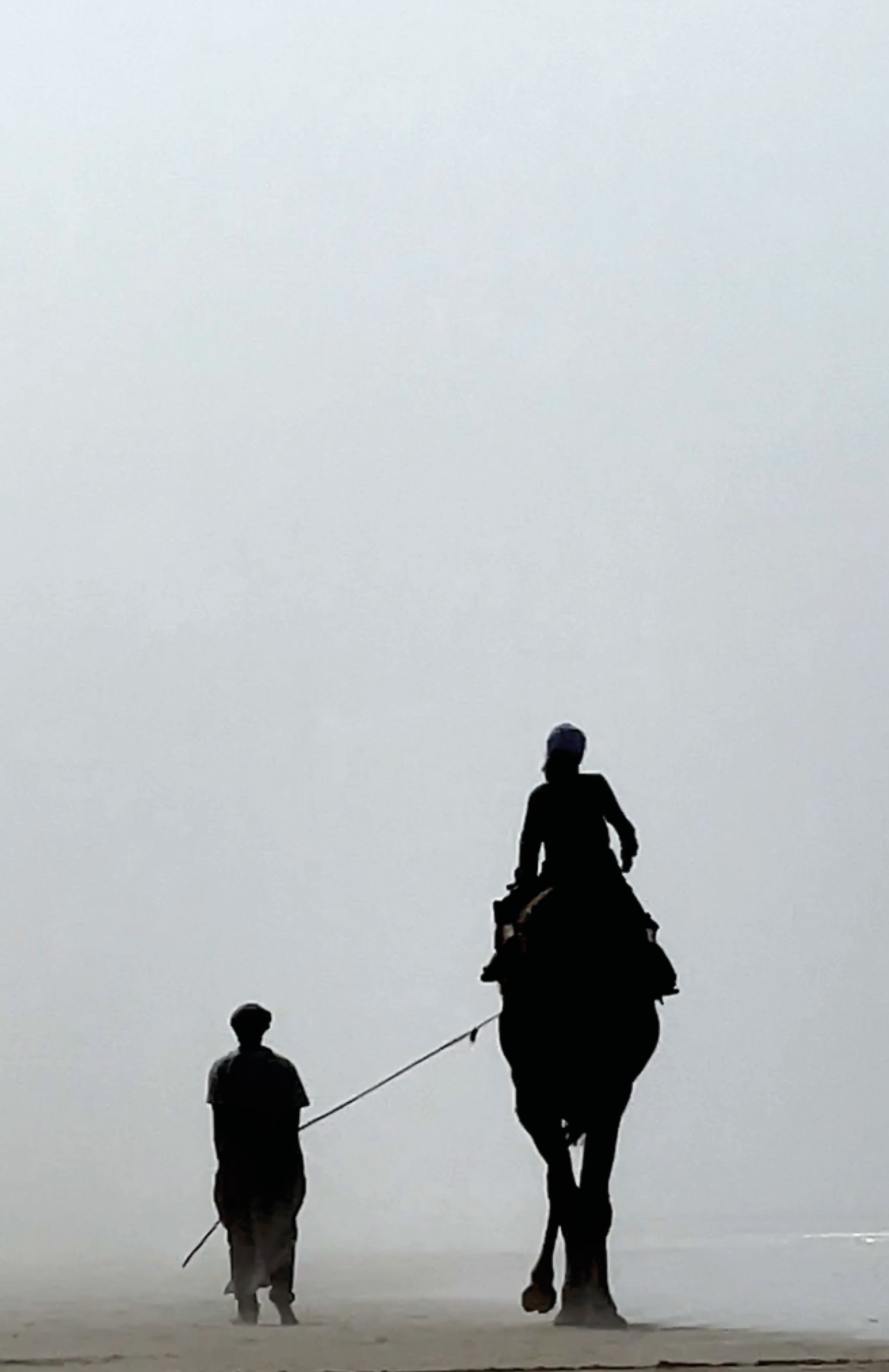 A person walks along the beach with a large horse carrying another person.