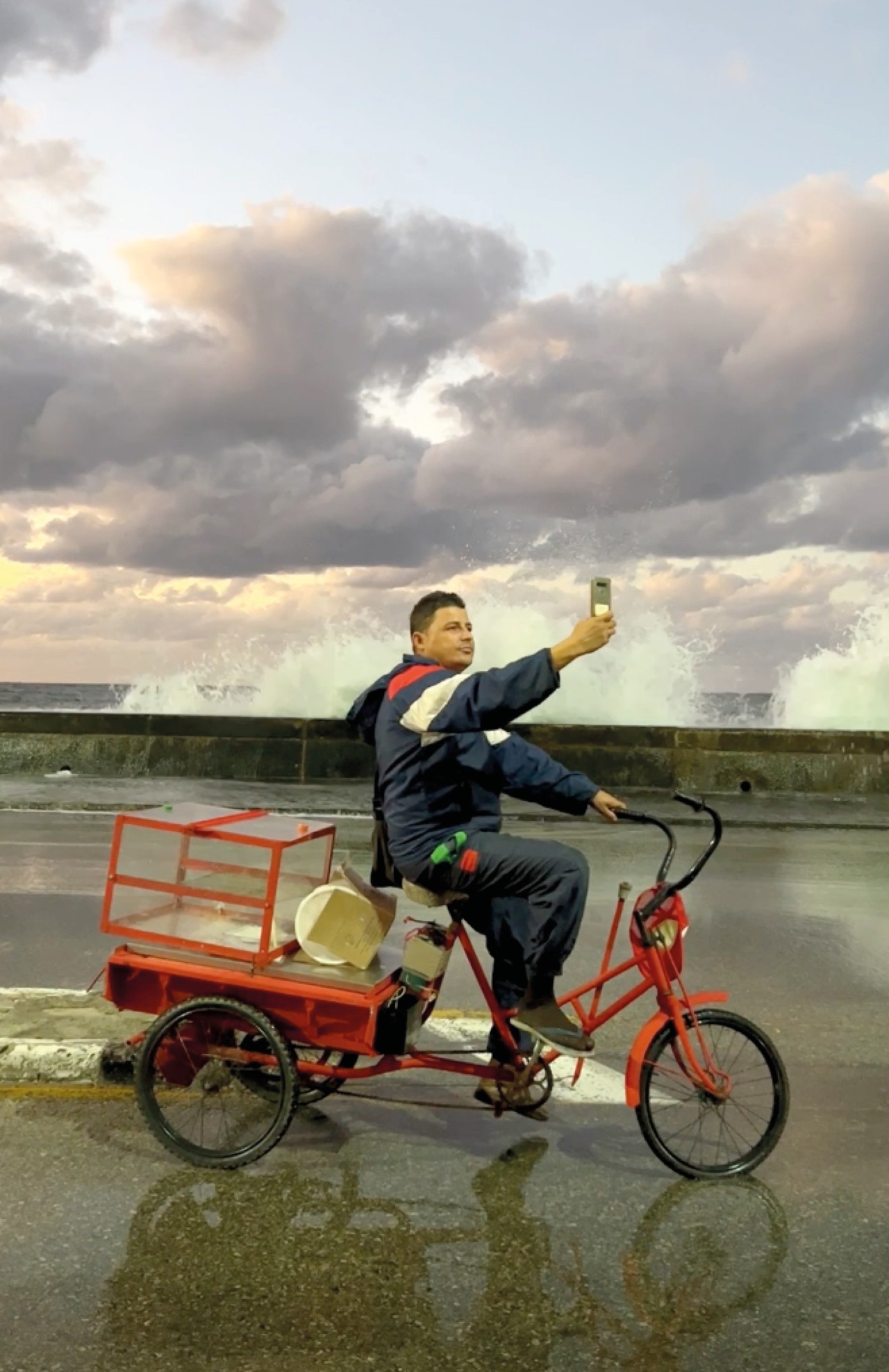 A person holds up a phone while riding a red bike along the shore.