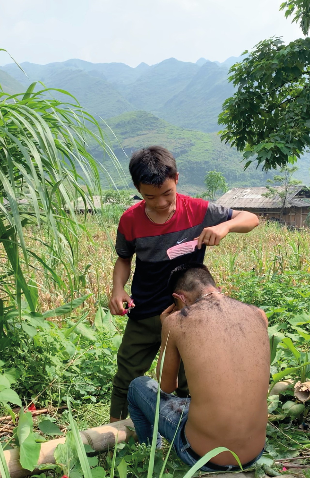 A young person combs the hair of another young person while standing in a grassy field with mountains in the background.