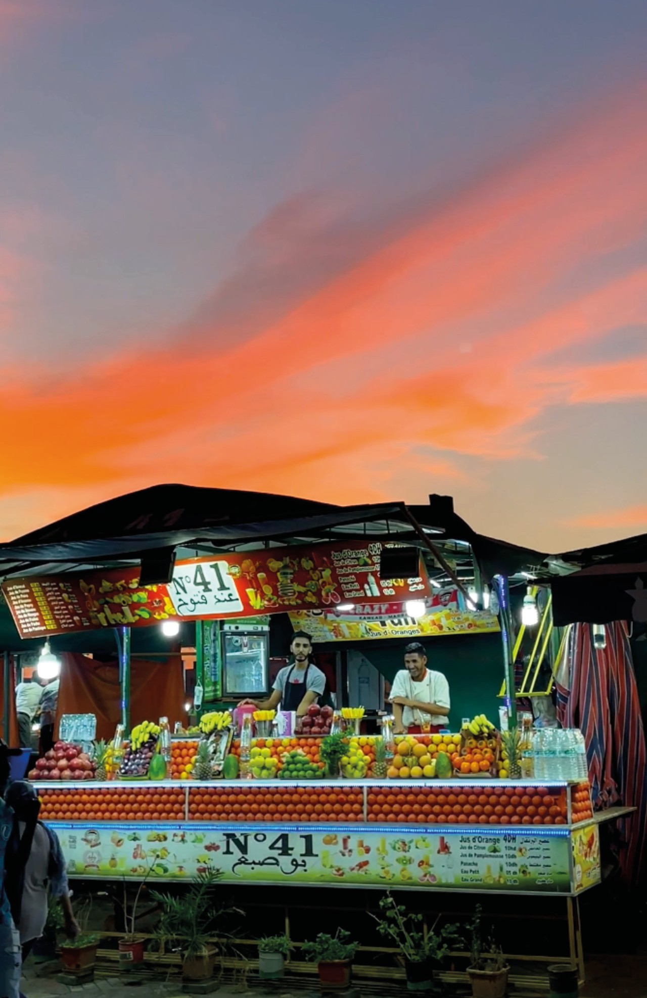 Two vendors stand at large fruit stand with the orange blue sky in the background.