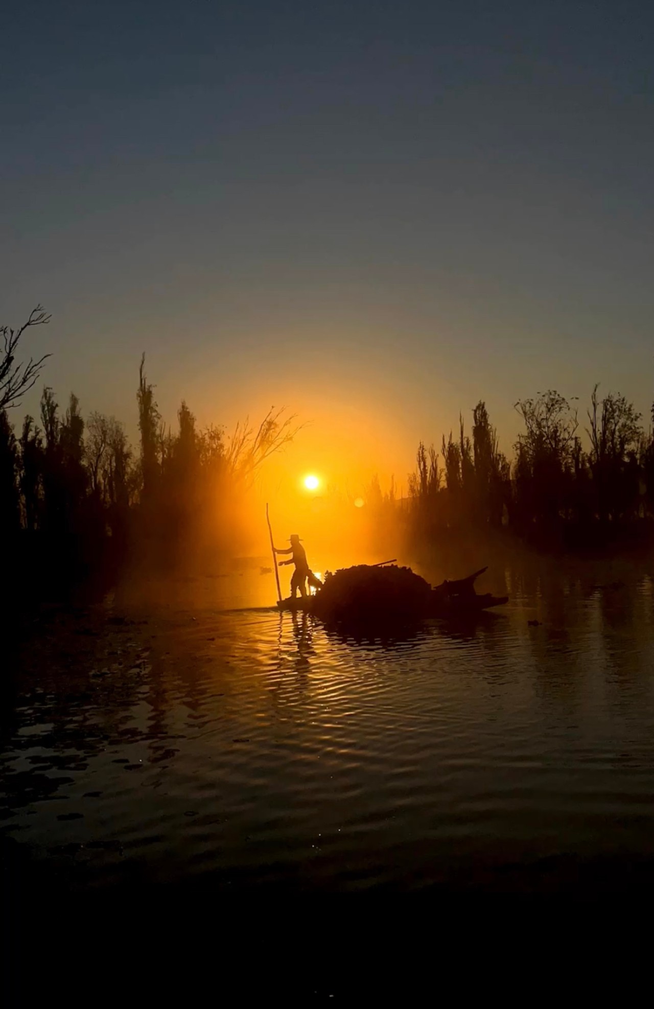 The sun sets over a small body of water where someone is standing on a rock.
