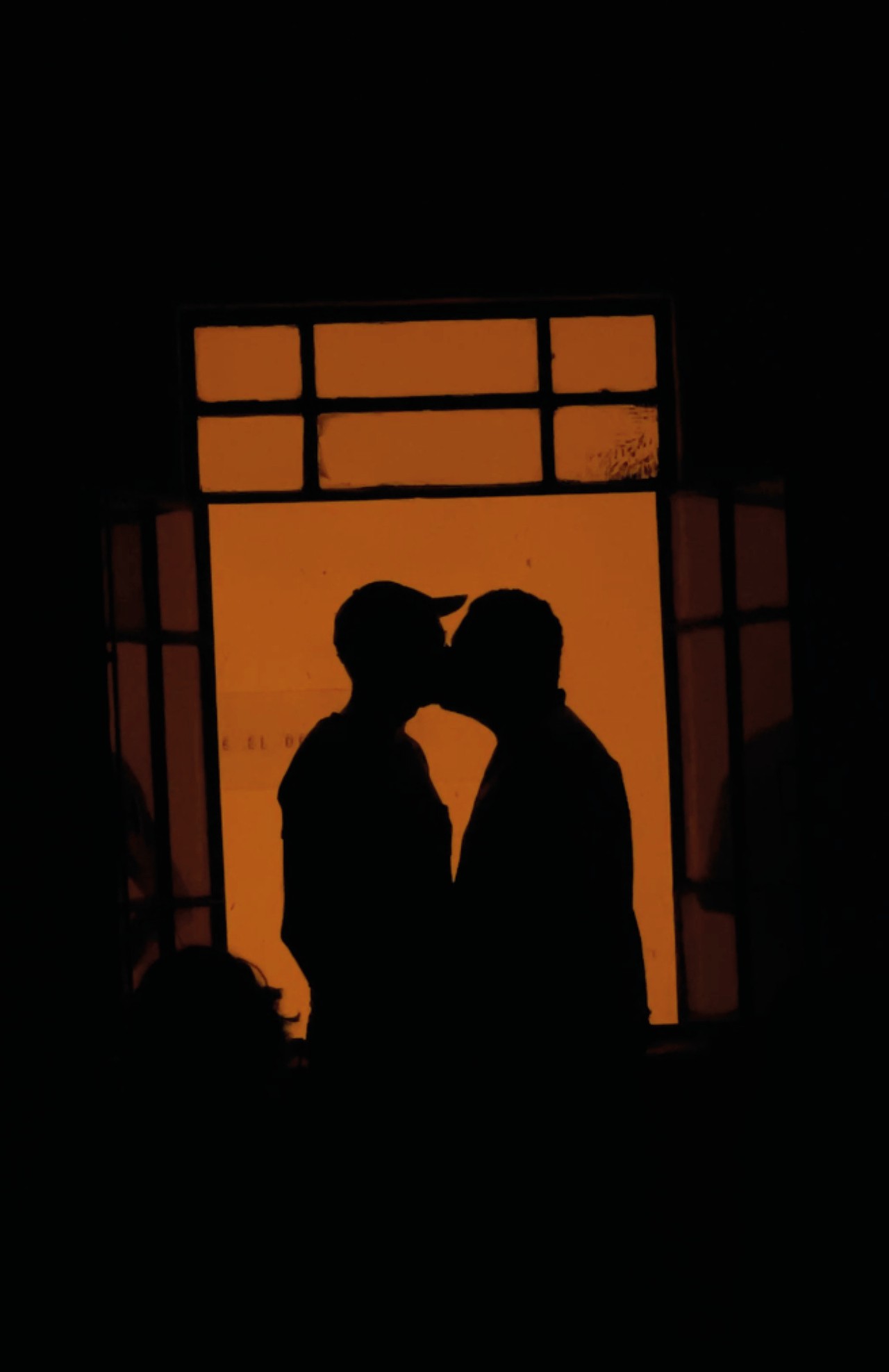 The silhouettes of two people embrace in a kiss through an orangey window.