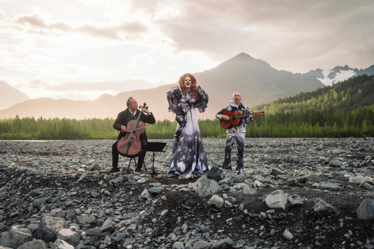 Pattie Gonia, Yo-Yo Ma and Quinn Christopherson perform while standing in a rocky clearing with mountains in the background.