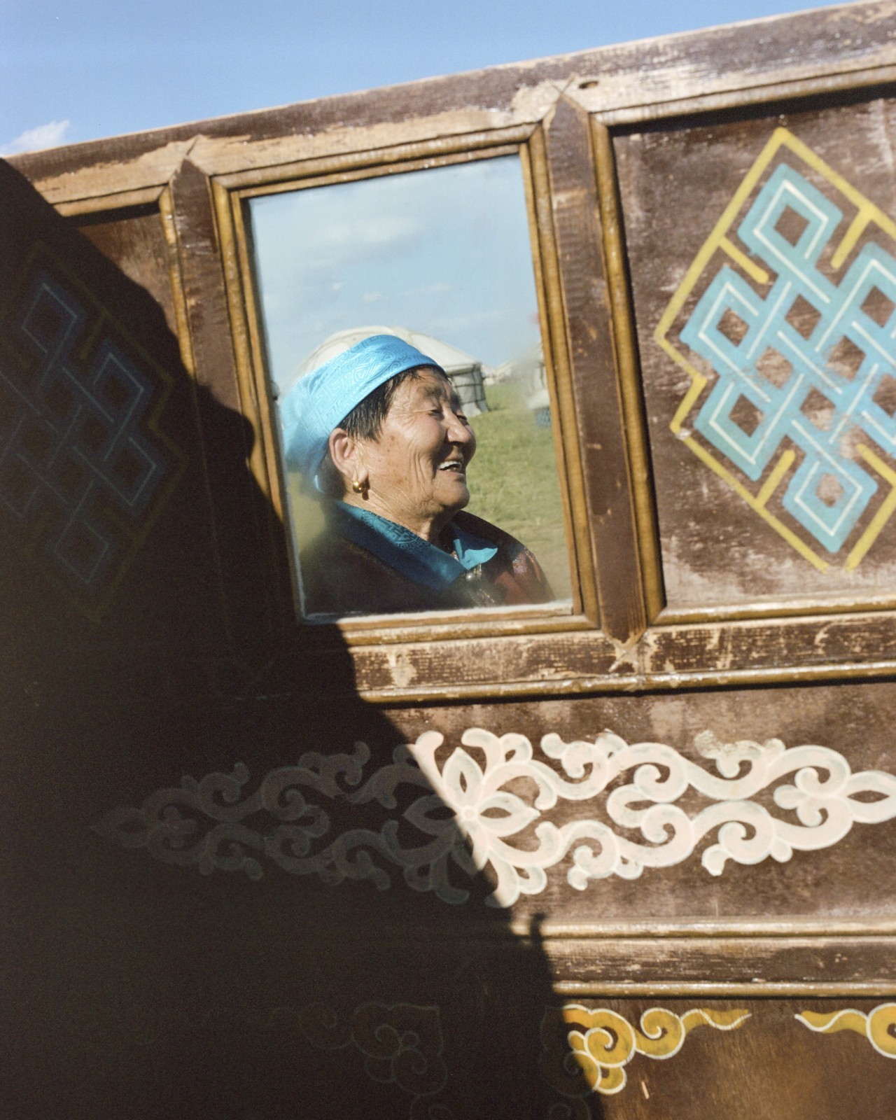 A Mongolian person laughs while standing behind a decorative brown wall with an open window.
