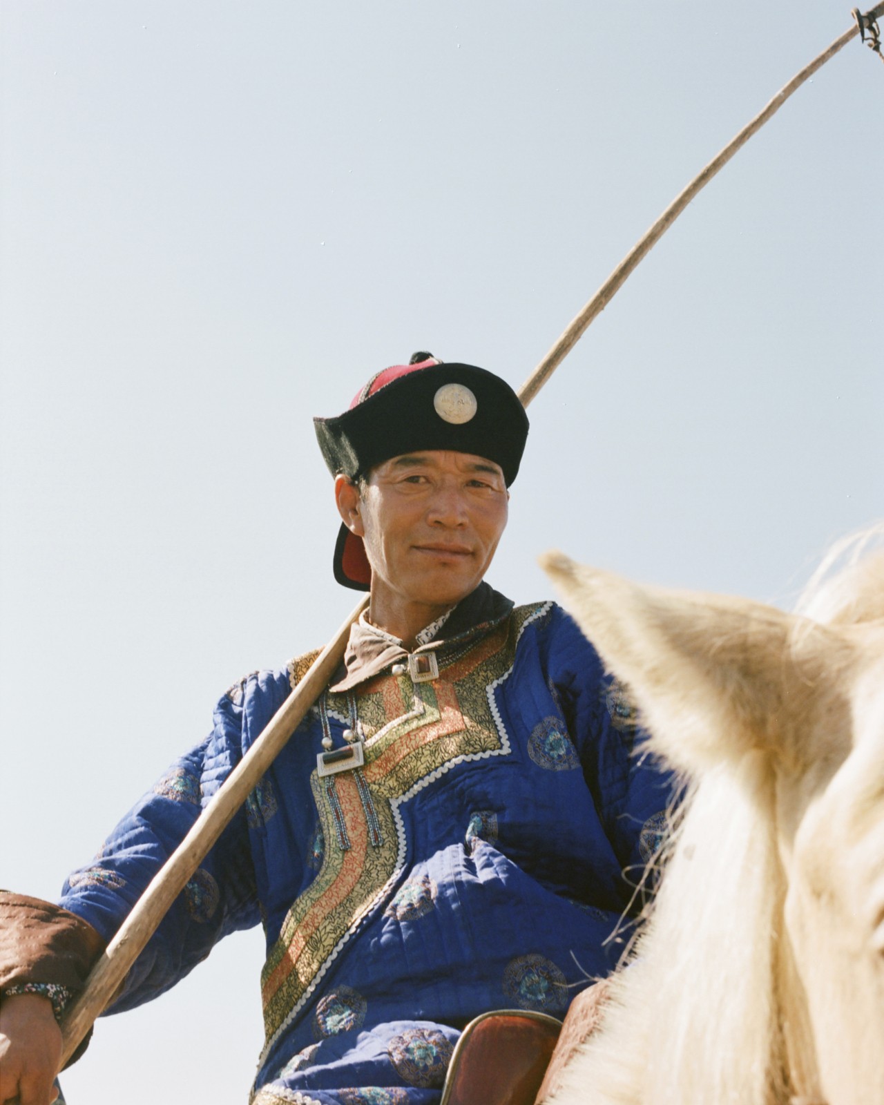 A mongolian person holding a long stick peers down from the top of their horse.