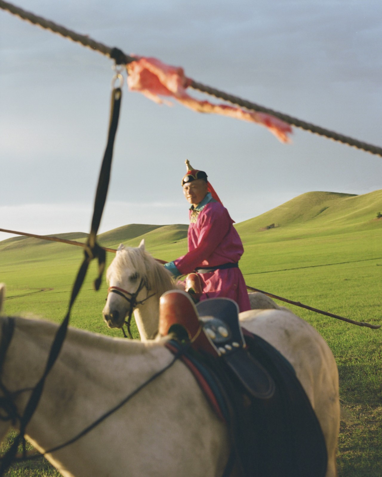 A Mongolian peron sits on their horse in a grassy mountainous field.