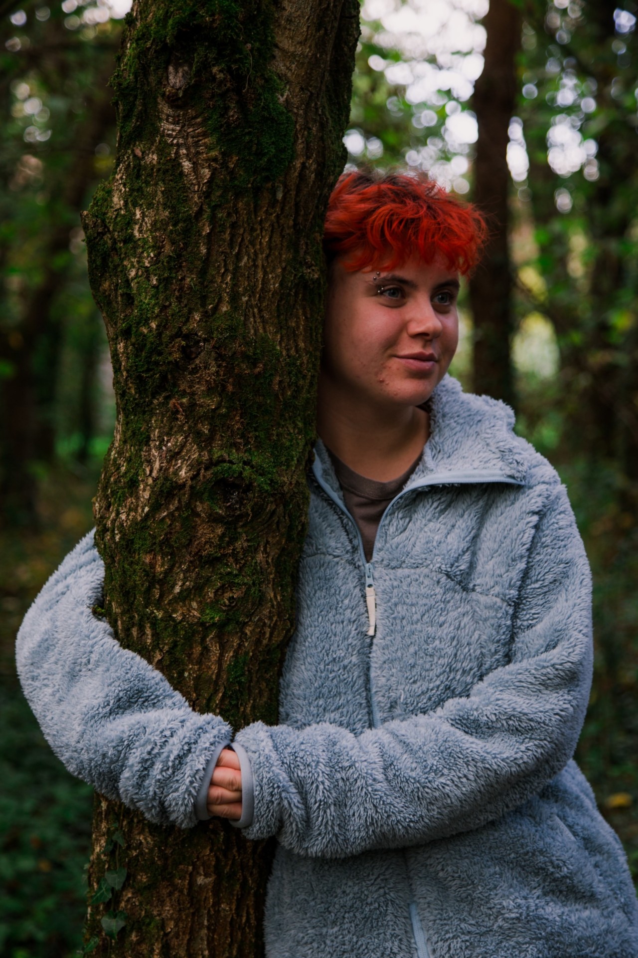 A young person with red hair wraps their arms around the trunk of a tree in the middle of the forest.