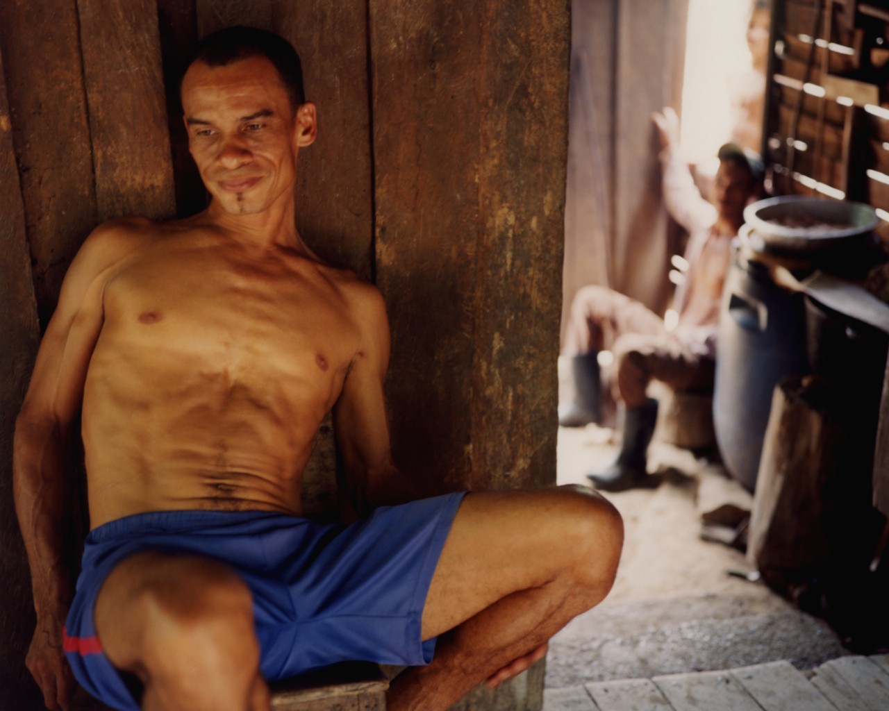 An older man wearing blue shorts sits leaning against a wooden wall inside a home.