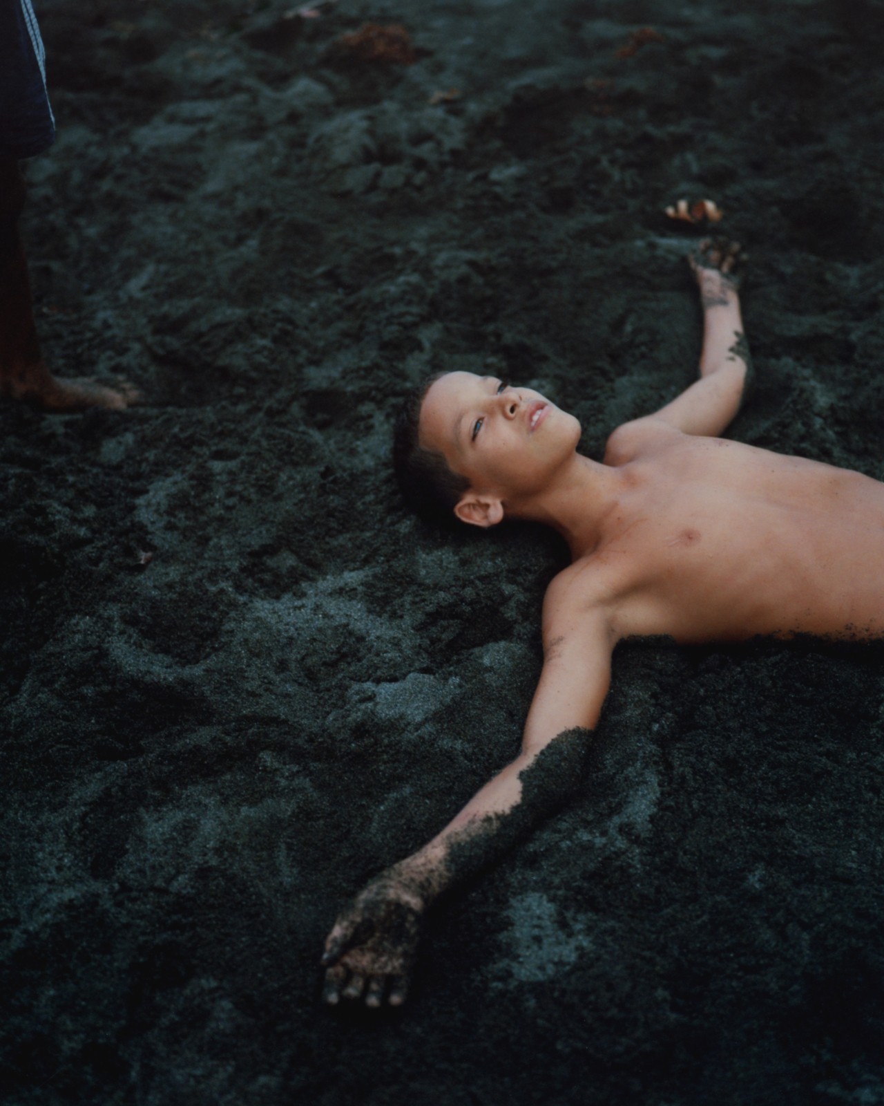 A child lies down in the charcoal sand with their arms outstretched.
